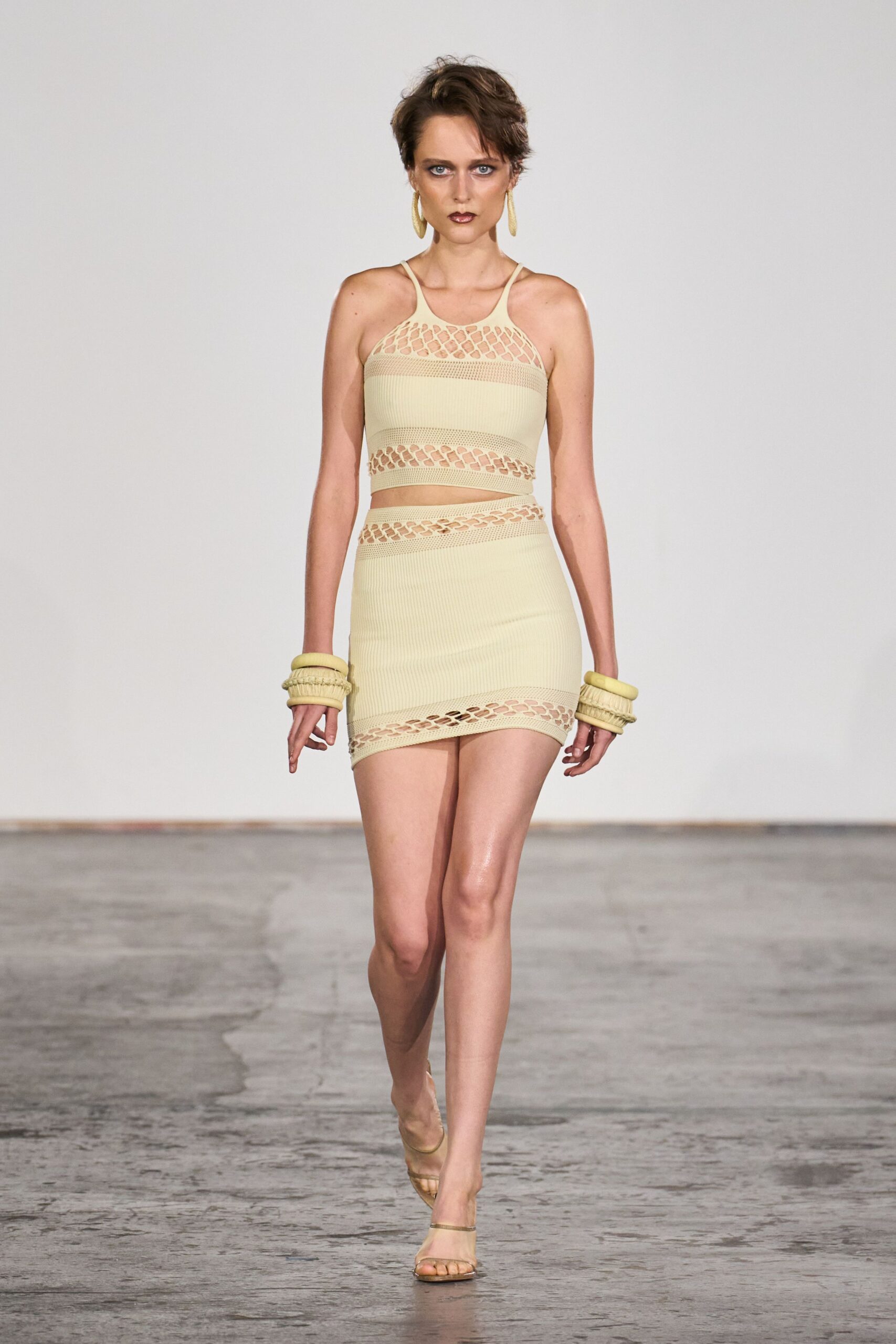 Buttercream rib knit crop top with open lattice at the chest. Matching ribbed miniskirt, hem band cut in the same linked openings. Stacked padded bangles in matching knit. Long oval drop earrings. Barely there thong sandals. Walking across a sunstruck hotel atrium before the pool crowd is awake.