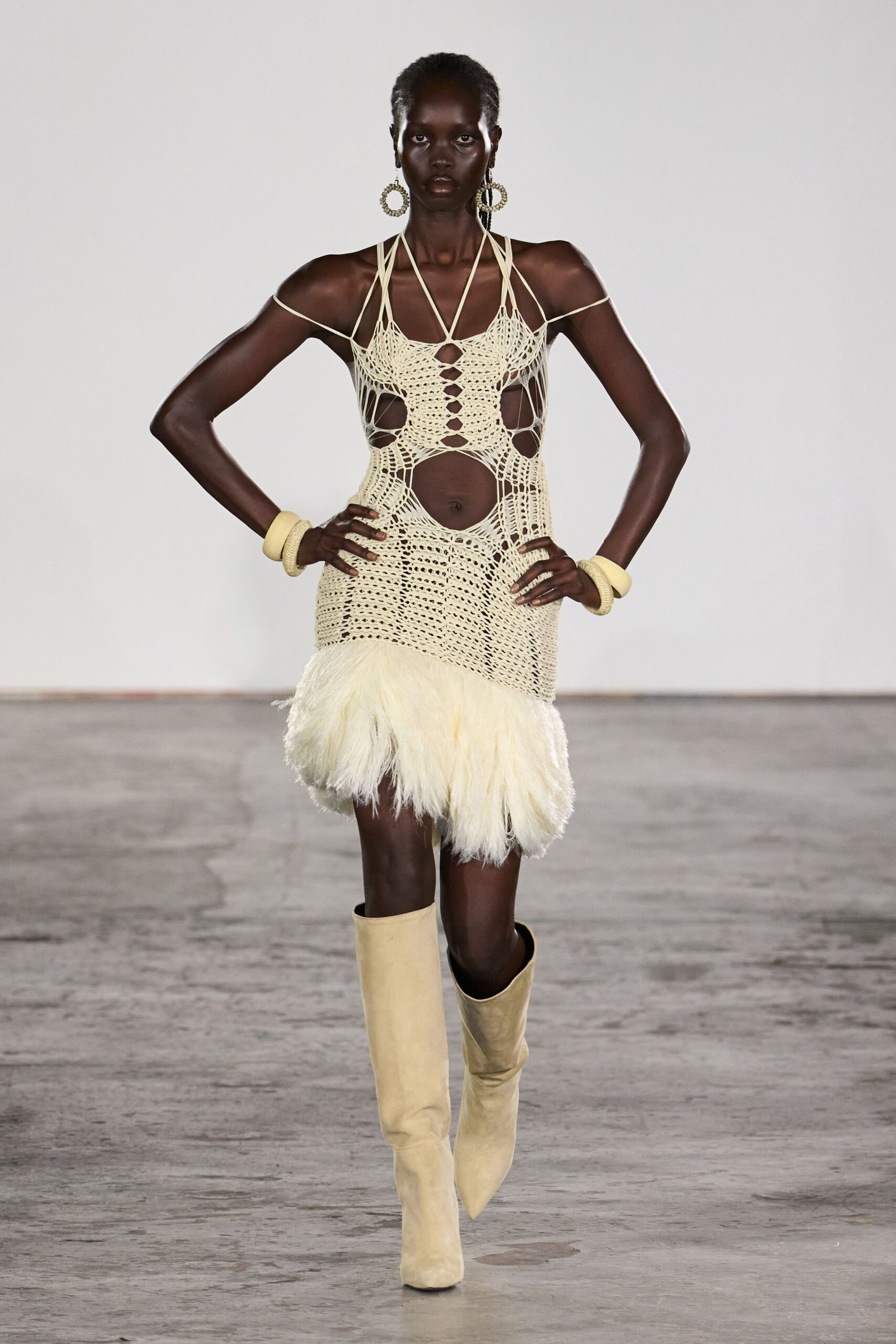 Large loop earrings beaded into uneven ivory rings. Openwork crochet minidress in cream cord, spiderwebbed across the torso. Long ivory hair fringe bursting from the dropped hem. Chunky butter bangles stacked at both wrists. Pale suede knee boots with sharply pointed toes. Walking from the beach bar before the floor has dried.