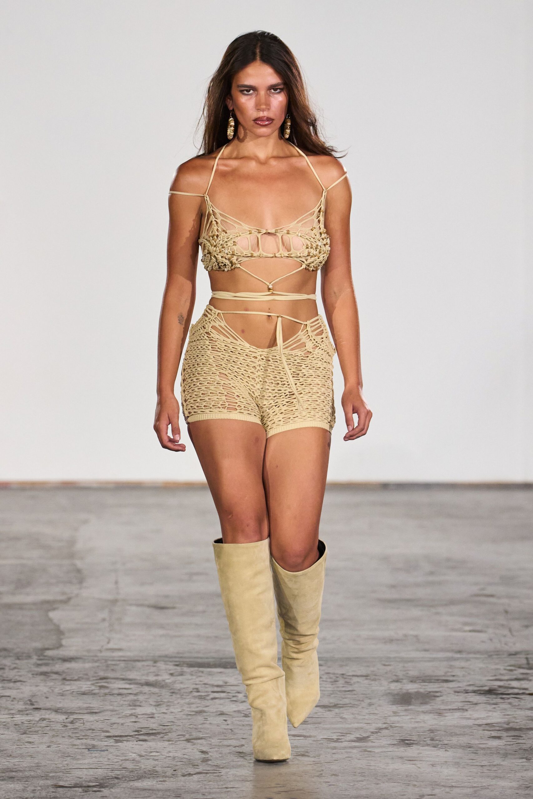Shell-drop earrings stacked in uneven cream discs. Sand crochet bralette, laddered cutouts and cords wrapping the bare waist. Openwork knit shorts with a high V waistband and dangling front tie. Pale suede knee boots with blunt pointed toes. Walking back from the beach bar after the boardwalk emptied.