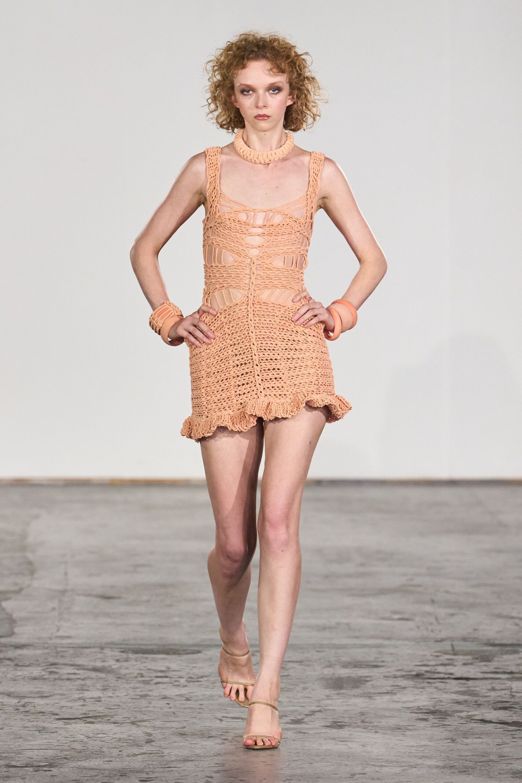 Chunky peach braided collar sitting close at the throat. Peach open-knit mini dress, laddered cutouts and a ruffled hem kick. Stacked peach resin bangles on both wrists. Barely there nude sandals with thin toe and ankle straps. Walking into a beachside hotel lobby still salty from the afternoon swim.