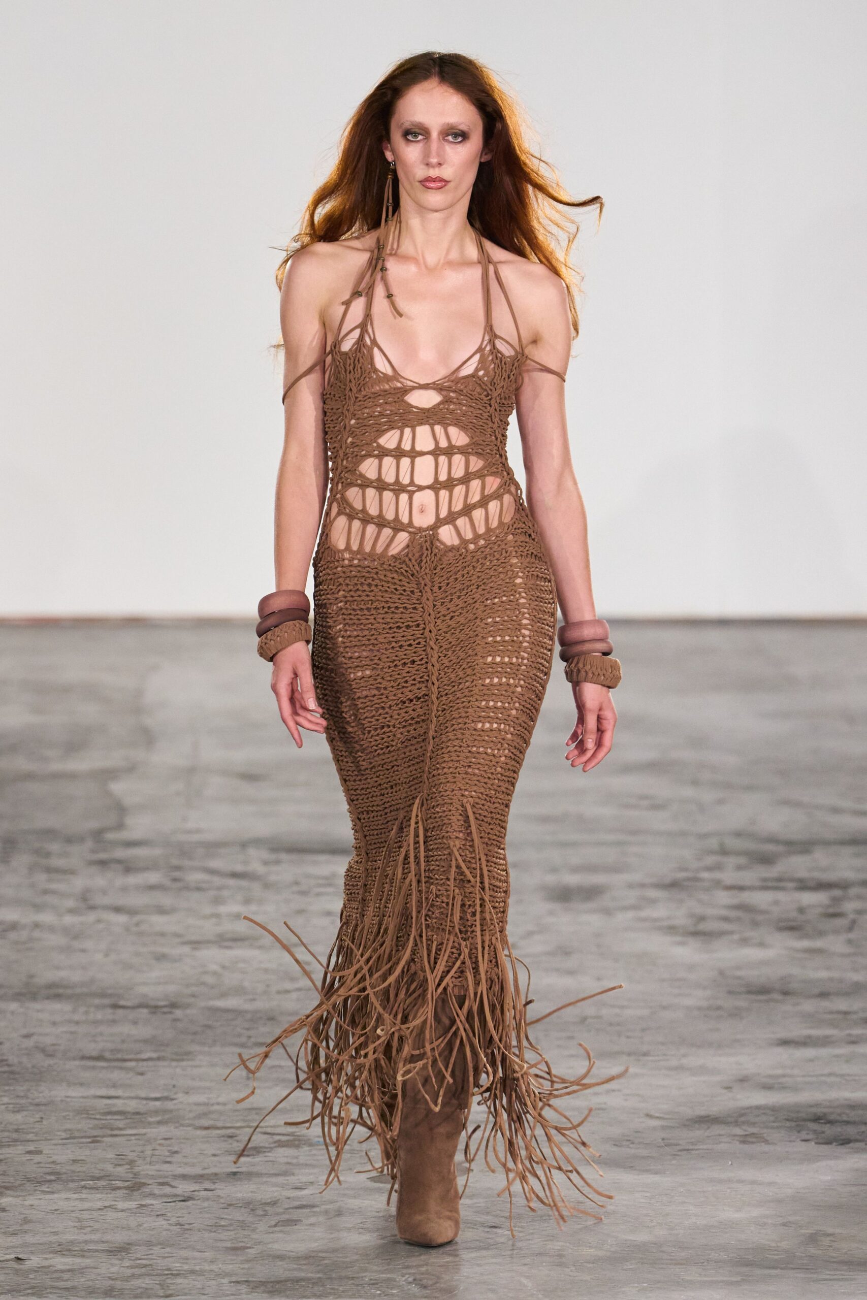 Taupe macrame halter column dress, torso opened into laddered cutouts. Stacked resin bangles over woven cuffs at both wrists. Single shoulder-grazing tassel earring with tiny bead spacers. Taupe suede boots buried under a whip of floor-sweeping fringe. For crossing a dry lake bed after the music has packed up.