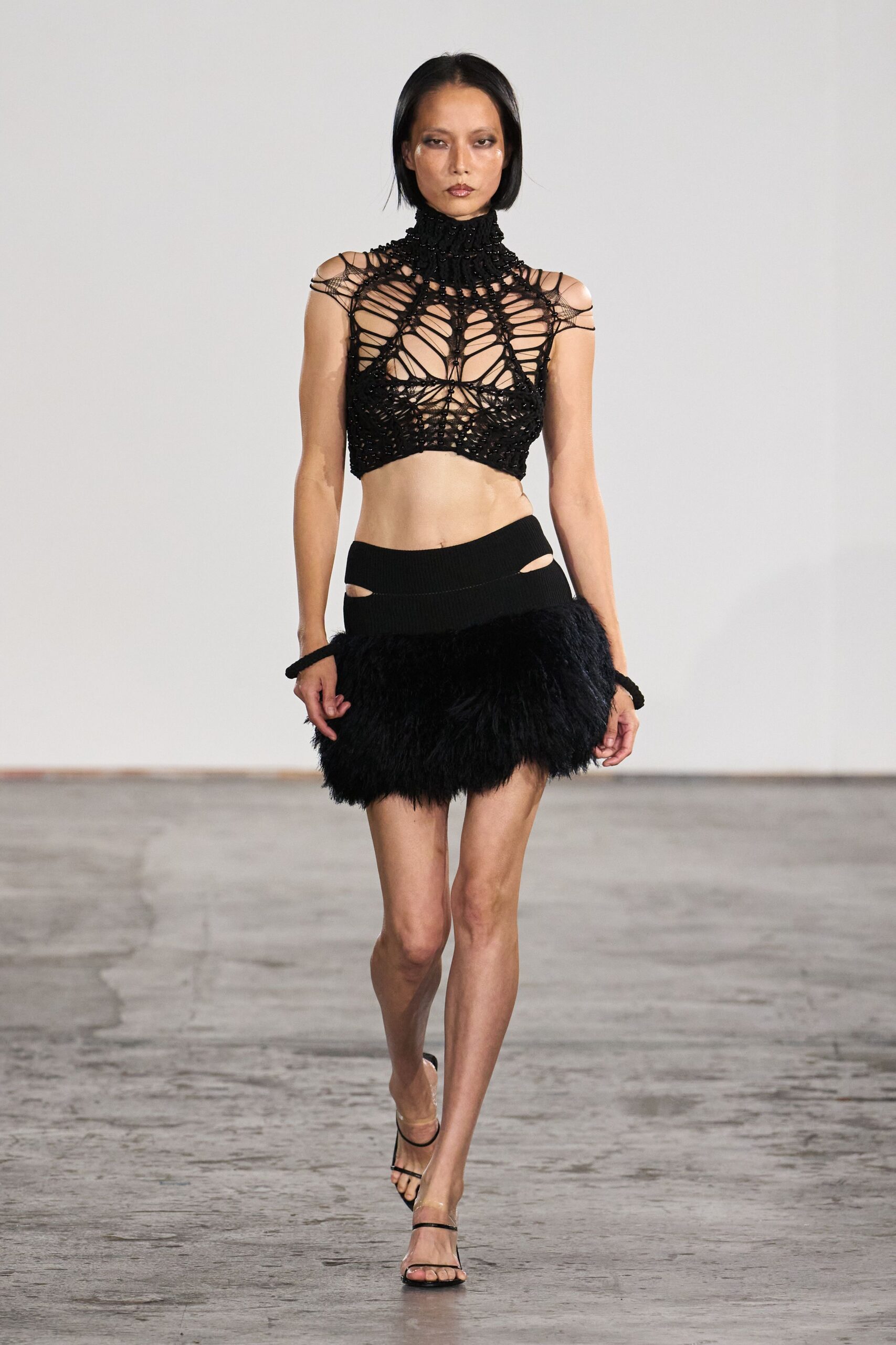 Black openwork halter, beaded cord lattice climbing into a ruched turtleneck. Low-slung ribbed mini with hip cutouts and a shaggy black feather hem. Bare black strap sandals. For arriving at the hotel bar after midnight, still carrying cold air.