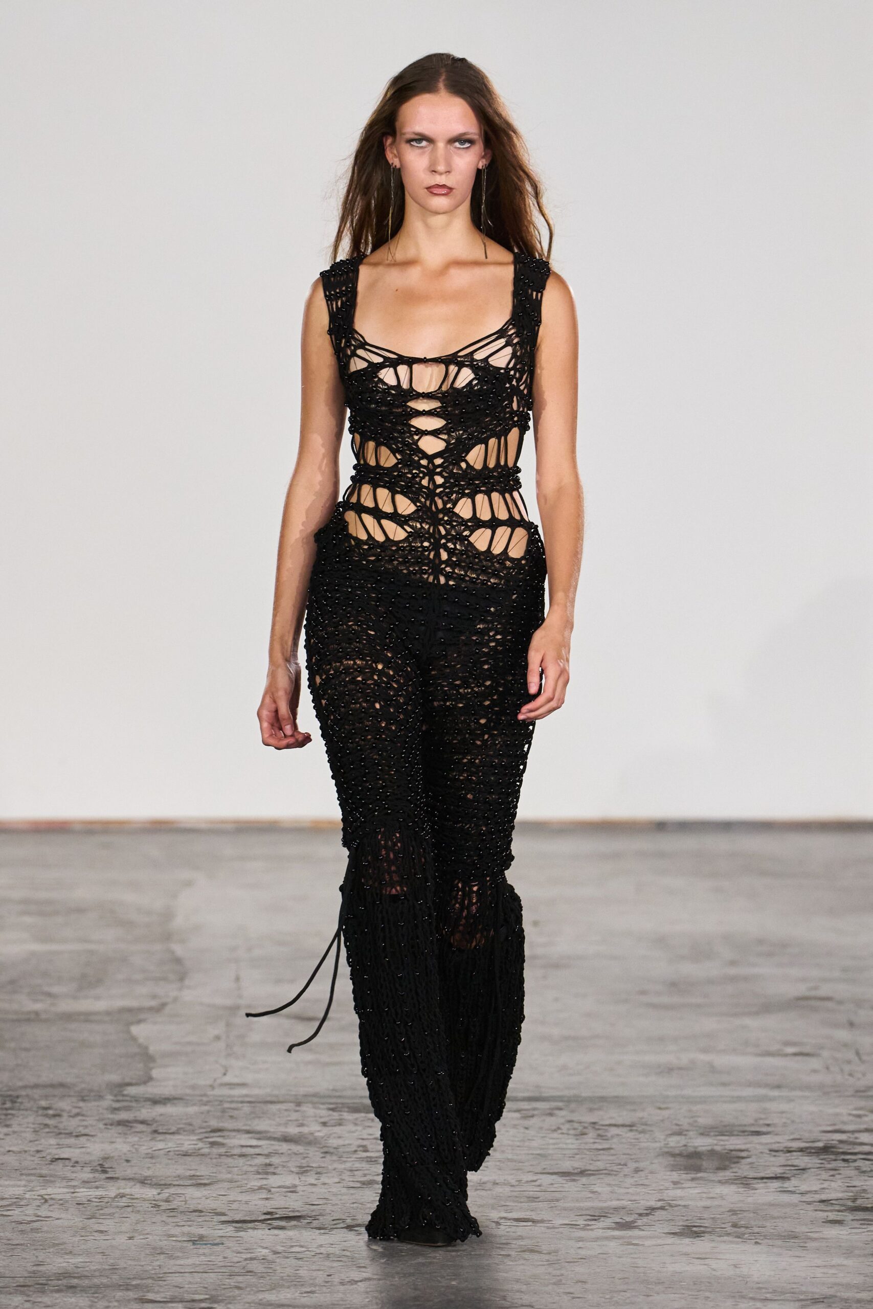 Black openwork cord jumpsuit, hand-knotted into netted cutouts and beaded ribs. Long wire-drop earrings grazing the collarbone. Walking home from the river after midnight, hem catching grit.