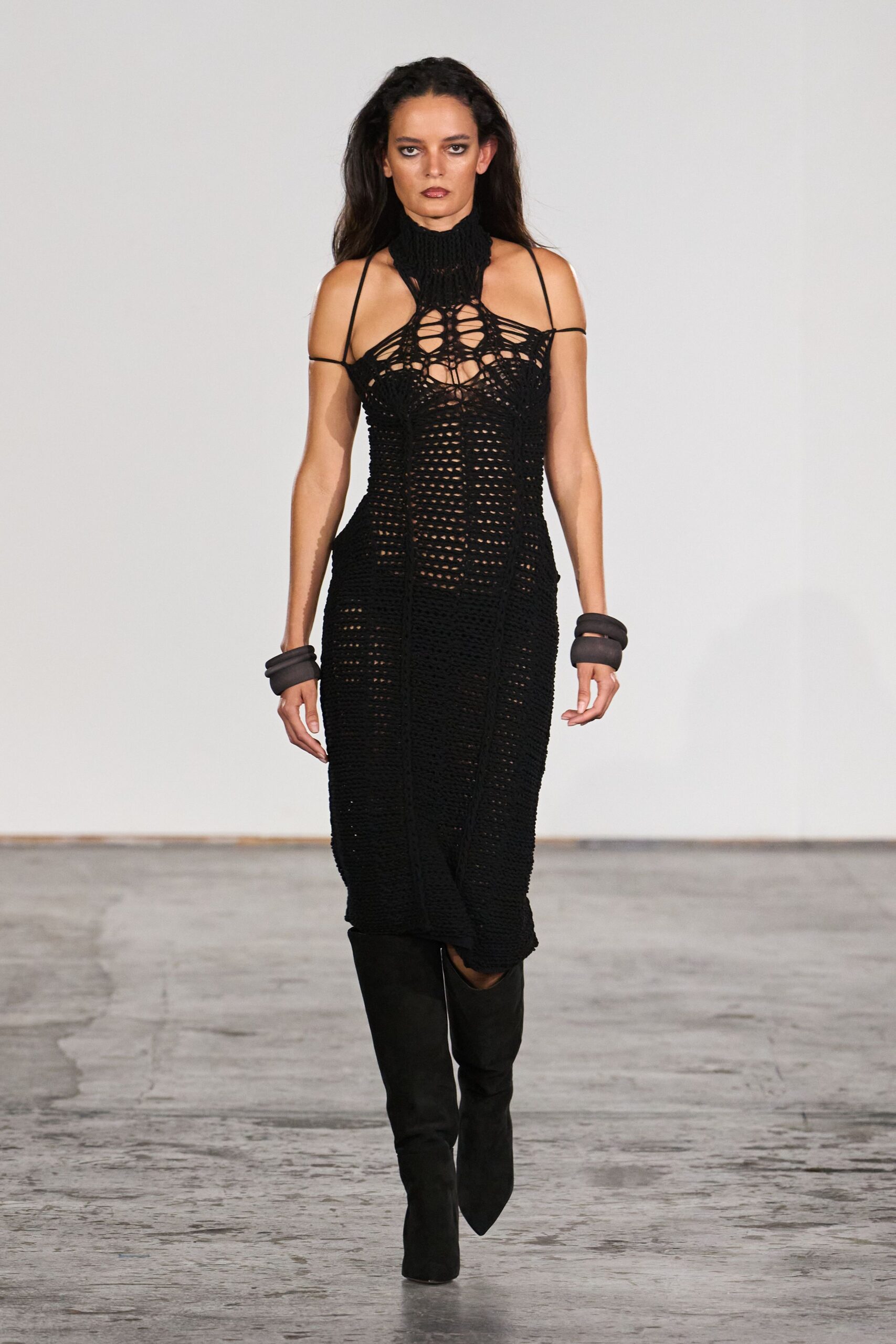 Black open-crochet column dress, spiderweb chest and ribbed neck cuff. Stacked charcoal padded rings at both wrists. Black knee boots with sharply pointed toes. For crossing a concrete lobby after midnight rain, still damp at the hem.