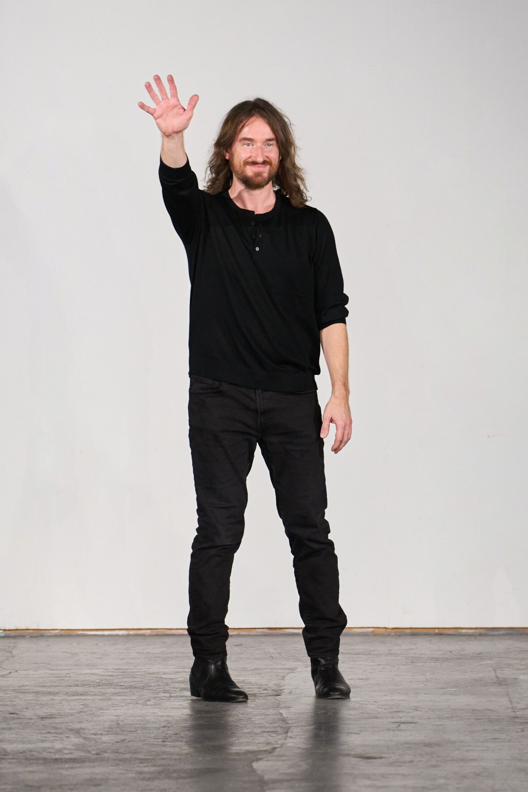 Black fine-knit henley, three-button placket and sleeves pushed unevenly up. Black slim jeans, faint whiskering across the upper thighs. Black leather ankle boots with softly creased vamps. Waving from the end of a warehouse presentation, concrete floor still warm.