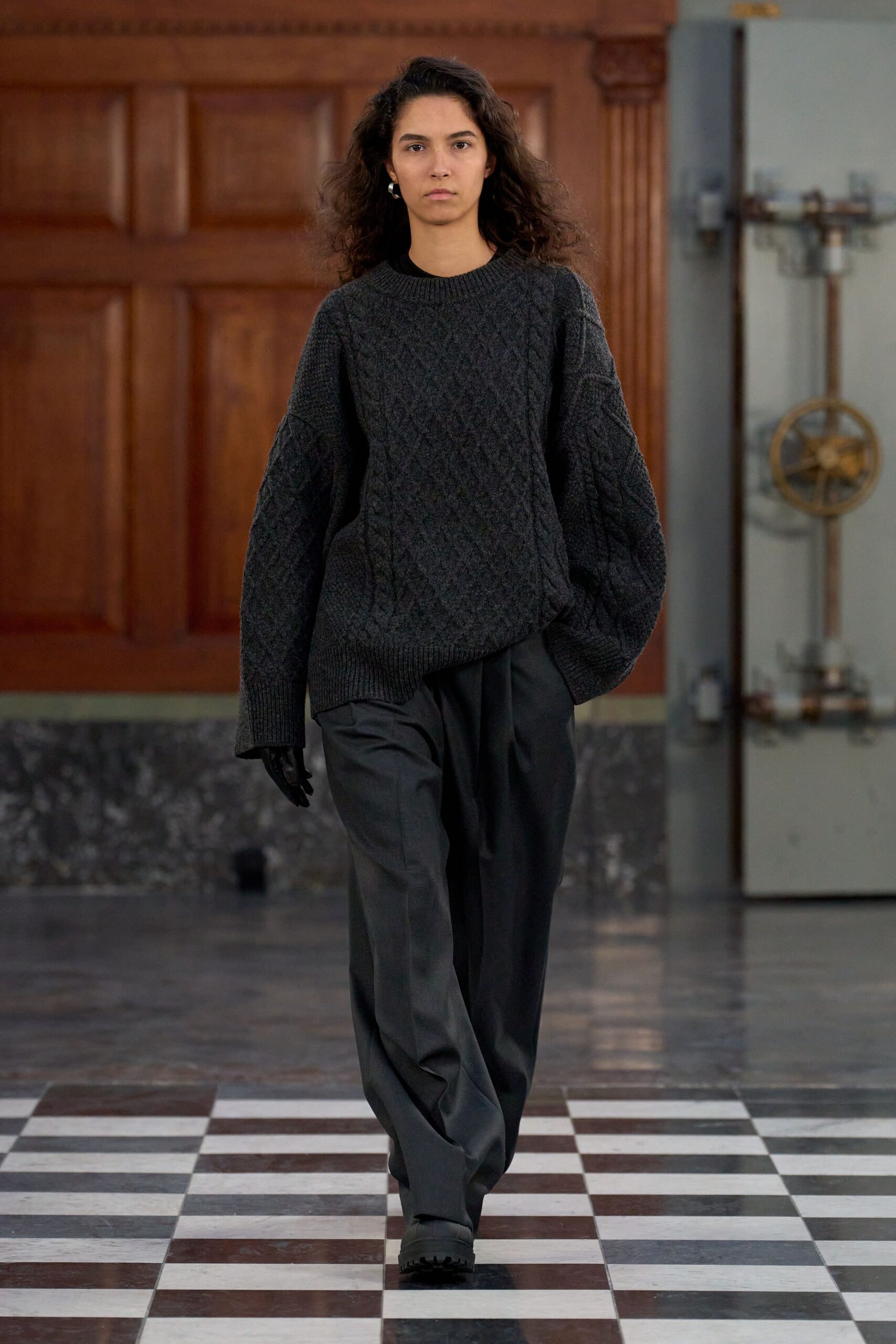 Charcoal cable-knit sweater with dropped shoulders and a hem dipping crooked. Dark wool trousers cut wide and long, breaking heavily at the shoe. Black leather gloves. Black lug-sole slip-ons. Crossing a draughty civic hall before the radiators have come on.