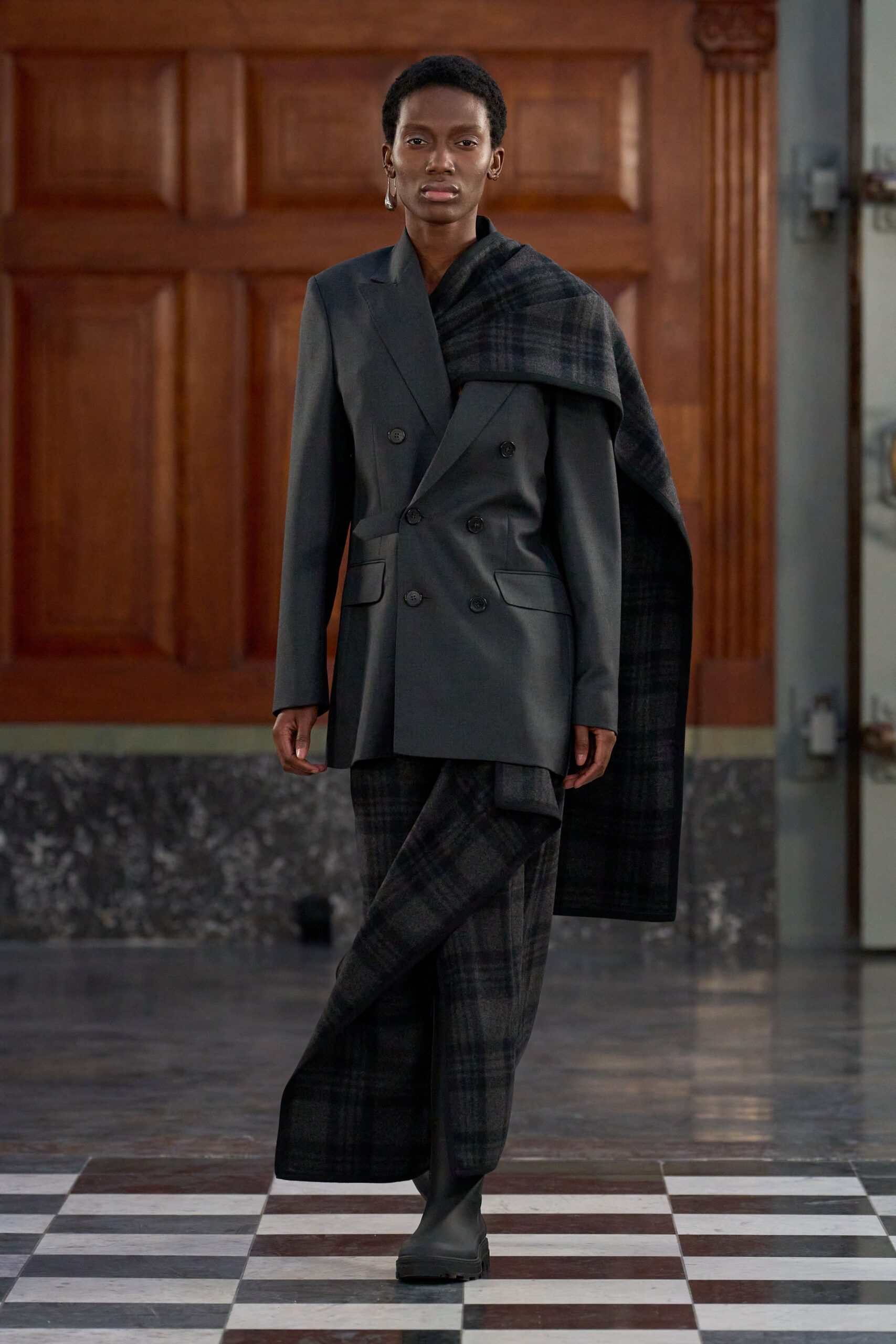 Charcoal double-breasted blazer in smooth suiting, one low button skewing the closure. Oversized charcoal plaid wool scarf slung heavily over one shoulder. Wide charcoal plaid trousers in soft brushed wool. Black rubber Chelsea boots with thick lug soles. Crossing a drafty municipal hall before the radiators catch up.