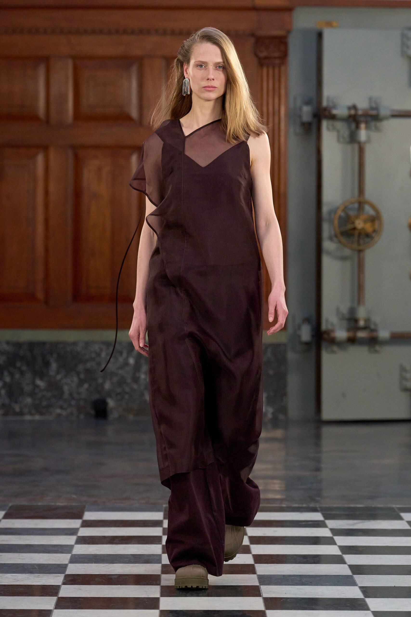 Single hammered silver slab earring brushing the jaw. Deep aubergine satin jumpsuit with a sheer organza panel and ruffled one-sleeve edge. Taupe rubber boots with thick tractor soles peeking under the hem. Crossing a drafty civic hall after rain, carrying the smell of wet stone.
