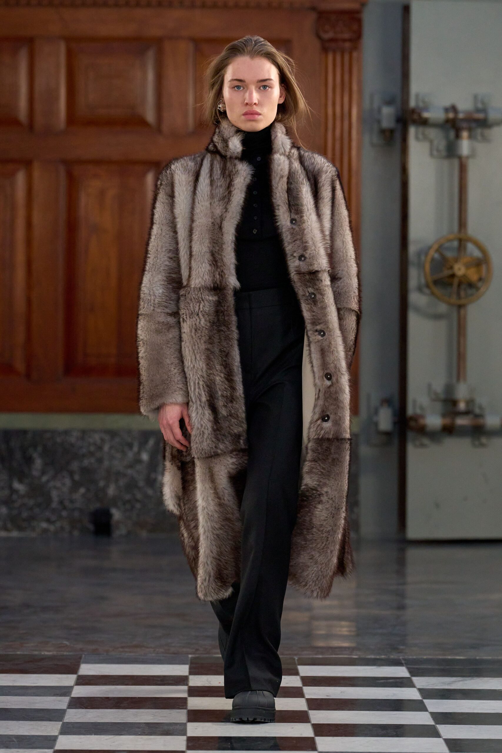 Long pieced fur coat in smoky taupe bands, dark buttons marching off-center. Black rib knit turtleneck with a short henley placket. Black tailored trousers falling straight from a sharp pressed crease. Rounded lug-sole boots in matte black leather. Crossing a drafty manor hallway before anyone has lit the fire.