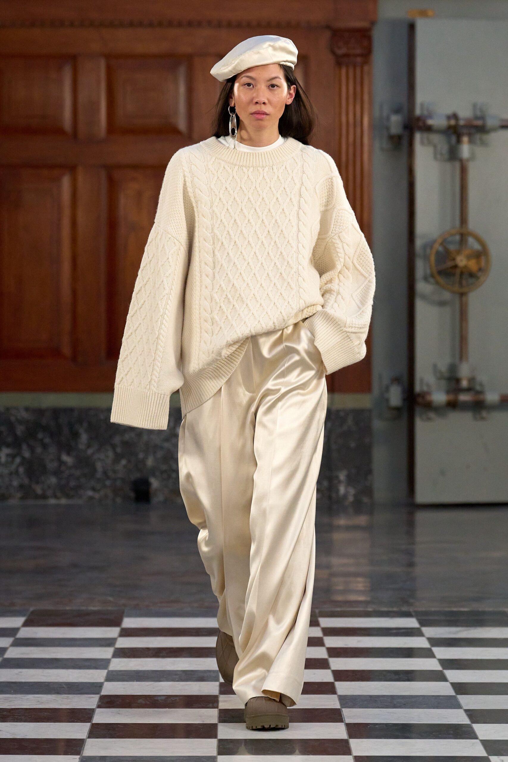 Cream beret tipped loosely to one side. Long silver link earrings. Ivory cable-knit sweater with balloon sleeves and a sharply ribbed hem. Champagne satin trousers, liquid shine pooling over the shoes. Taupe lug-sole boots. Crossing a museum atrium before the radiators have warmed up.