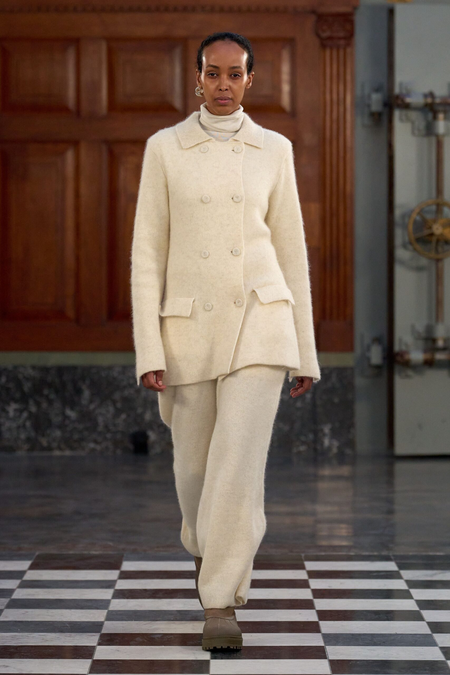Cream jersey turtleneck snug at the throat. Ivory brushed-wool double-breasted jacket, one flap pocket tipping outward. Matching wide-leg knit trousers with a soft, clouded surface. Taupe leather ankle boots with thick lug soles. Crossing a draughty manor hall before the radiators wake up.