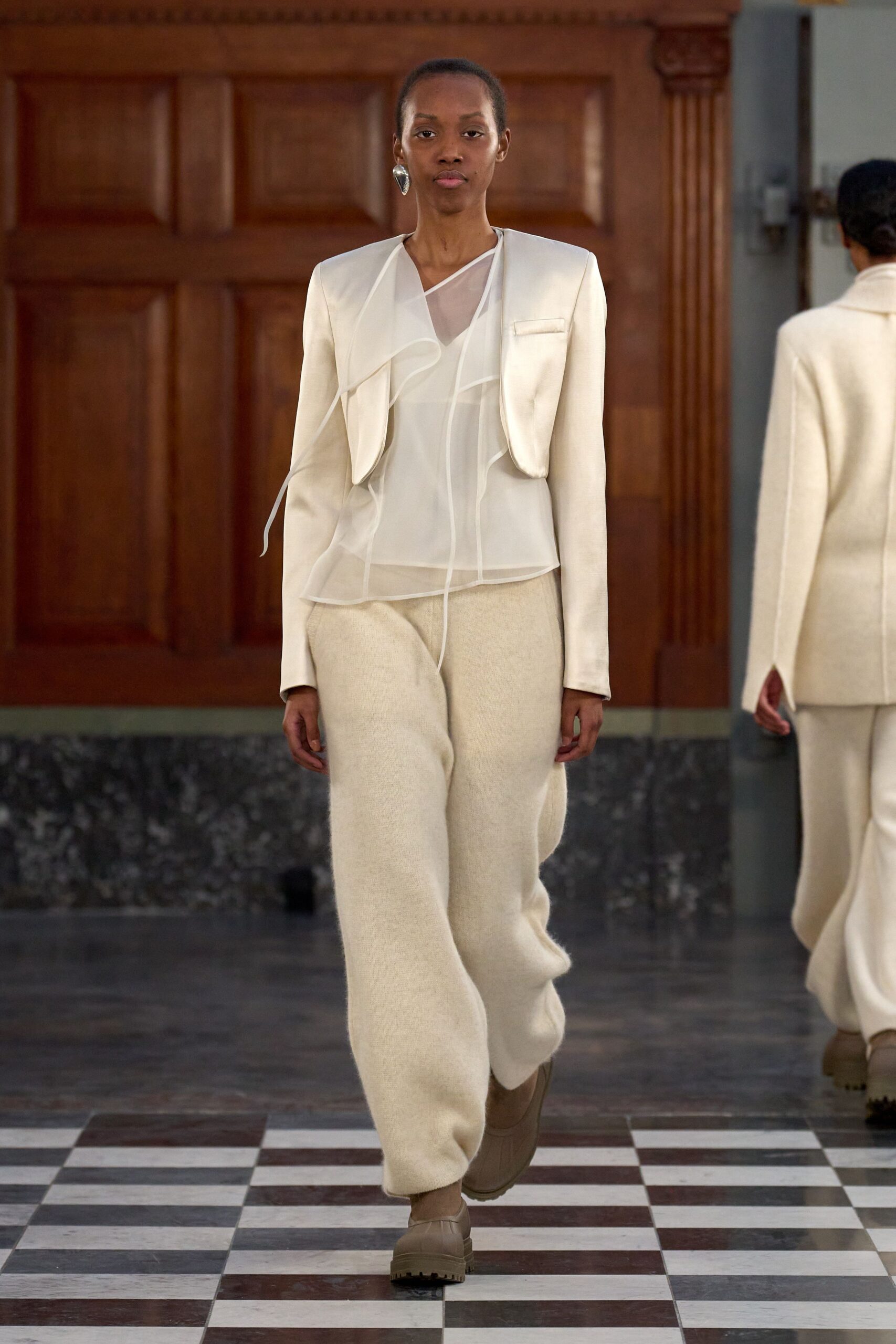 Single hammered silver drop earring. Ivory cropped jacket with curved front panels and no fastening. Sheer organza blouse, asymmetric neckline and loose piped ruffles. Cream brushed-wool trousers, cut full and soft through the leg. Taupe lug-sole slip-ons. Crossing a courthouse lobby before the heating has come on.