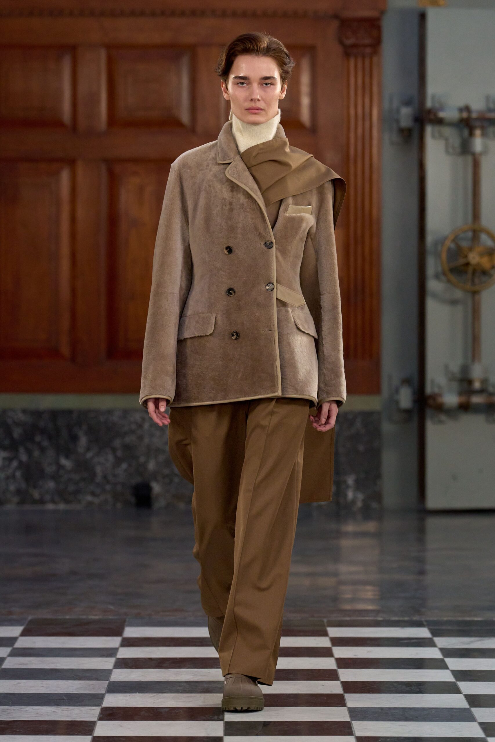Cream rib-knit turtleneck pulled high under the jaw. Taupe shearling double-breasted jacket, one shoulder wrapped in khaki suiting. Tobacco straight trousers with a pressed front crease. Taupe rubber boots with deep lug soles. Crossing a drafty municipal hall before the heating kicks in.