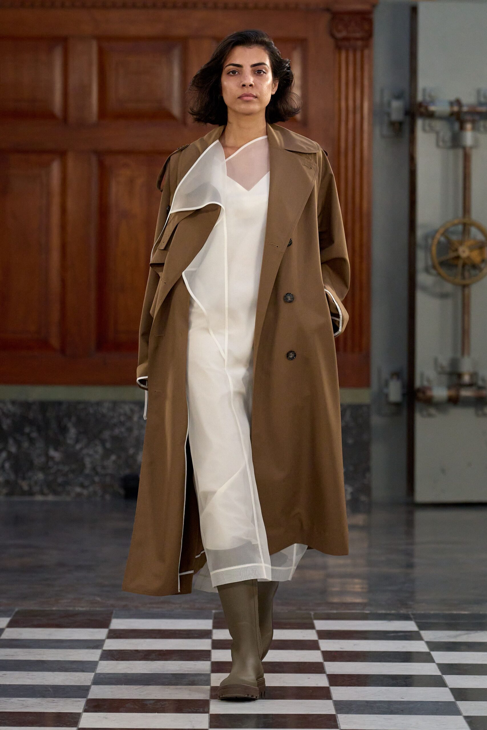 Tobacco trench with broad lapels and dark horn buttons. Ivory slip overlaid with sheer organza, white binding tracing a loose spiral. Olive knee boots in smooth rubbery matte finish. Crossing a municipal hall floor before the rain starts again.