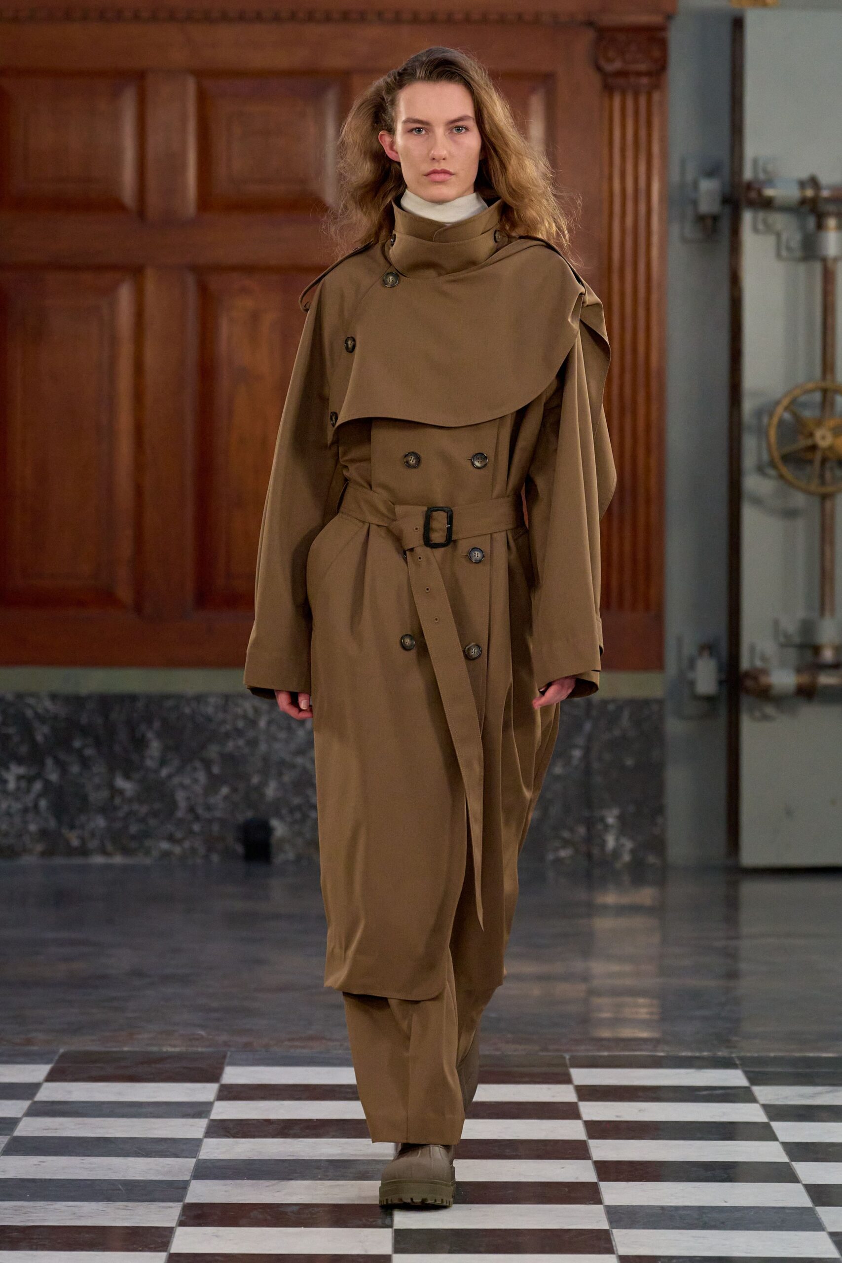 White jersey turtleneck tight to the throat. Taupe double-breasted trench with a high wrapped storm flap and dangling belt end. Matching straight trousers in the same dry twill. Sand ankle boots on thick lug soles. Crossing a municipal hall floor still wet from mopping.
