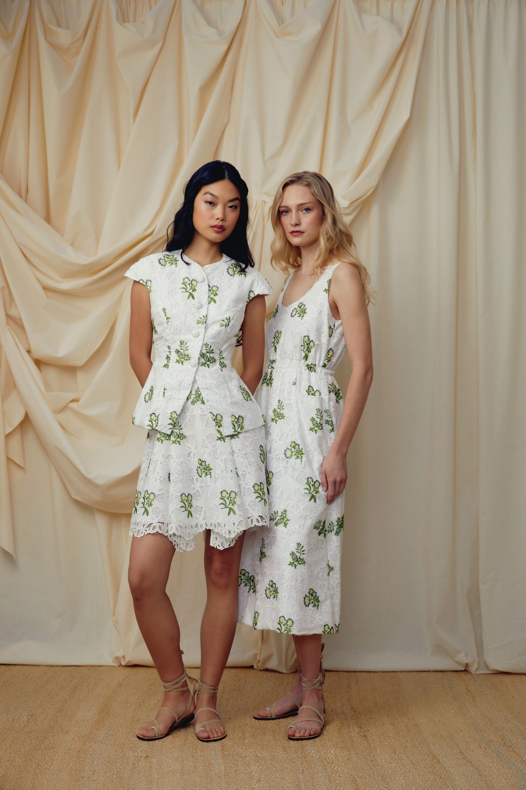 White floral eyelet top with padded cap sleeves and covered buttons. Matching mini skirt, scalloped lace hem peeking below the peplum. Barely there wrap sandals in pale beige leather. Garden wedding lawn gone soft after an afternoon sprinkler.