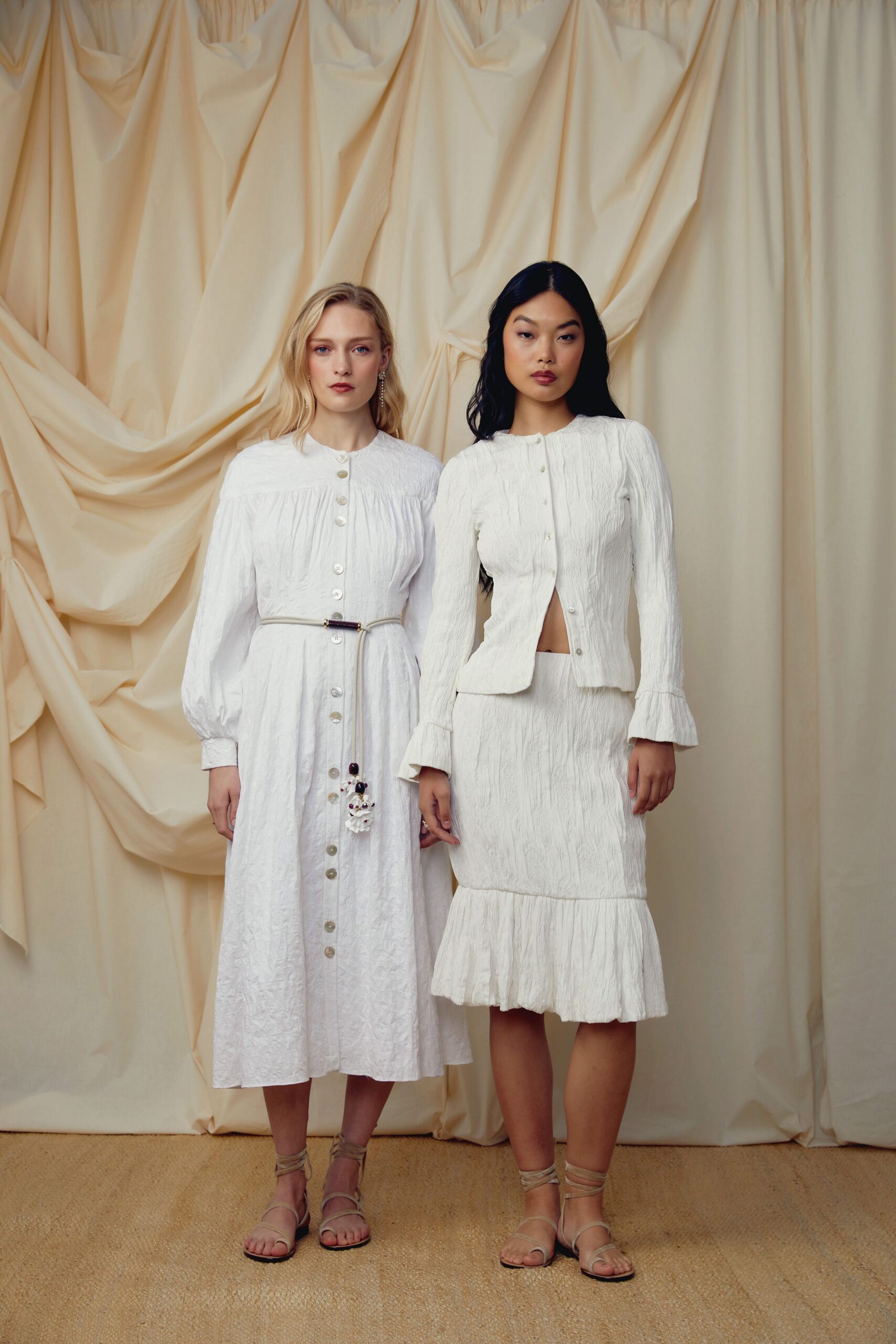 White crinkled knit cardigan, pearl buttons pulling open at the midriff. Matching crinkled midi skirt with a dropped flounce hem. Pale wraparound sandals laced up the ankles. Garden vows moved indoors because the grass never dried.