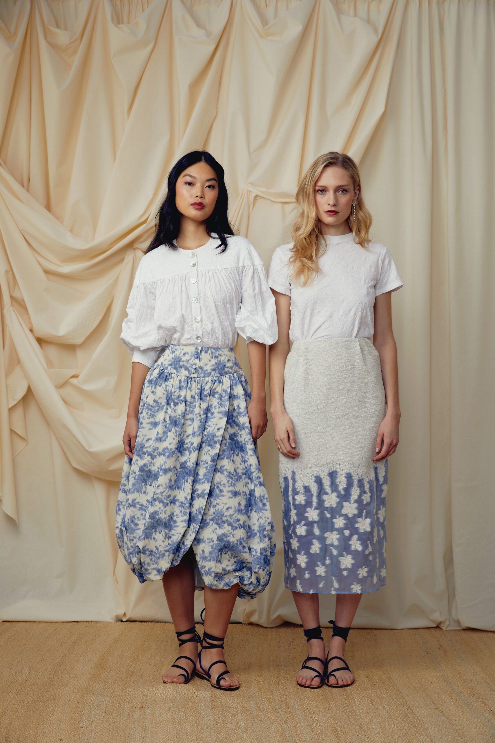 White cotton blouse with puffed sleeves and a row of pearl buttons. Blue toile skirt ballooned at the hem, one pleat falling deeper front center. Black wraparound sandals. Garden wedding brunch after the tables have already been cleared.