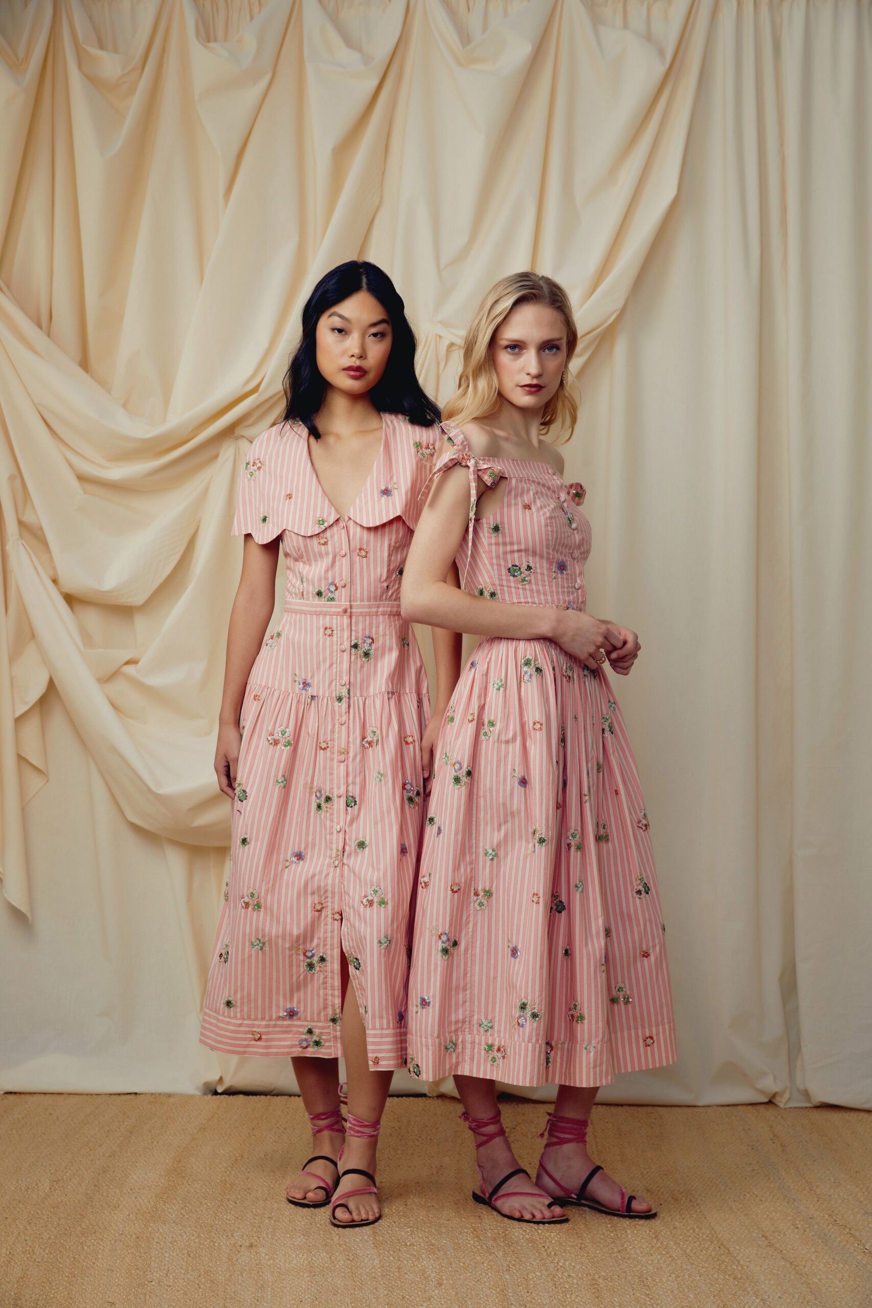Pink-and-white striped cotton dress with tiny embroidered flowers and a scalloped collar. Black flat sandals laced up the ankle in bright pink ties. Garden wedding morning, before anyone has found the cake knife.