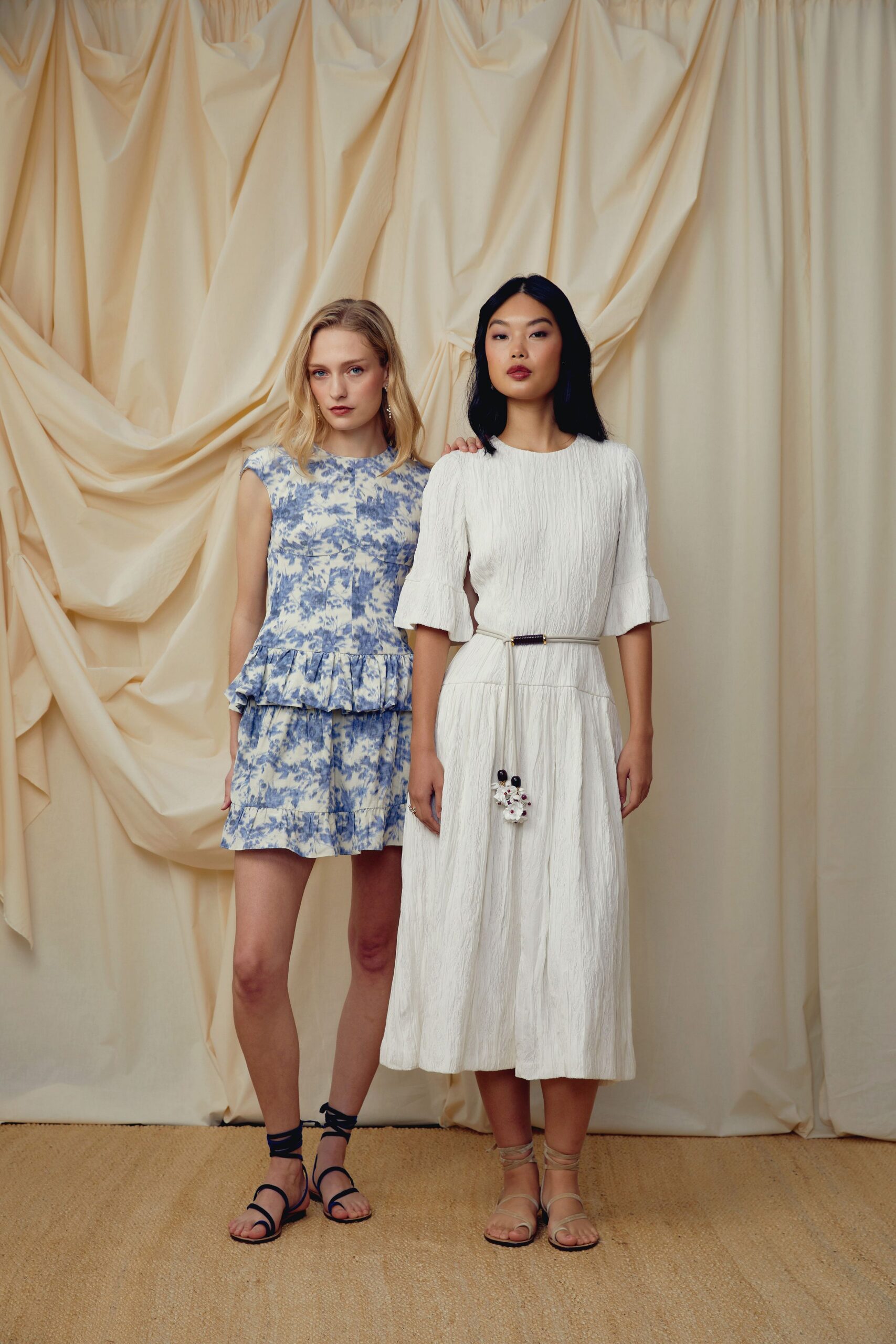 White crinkled midi dress with flounced elbow sleeves and a dropped waist seam. Thin cream cord belt with a tiny clustered bead tassel at center. Pale wraparound sandals laced up the ankles. Garden vows on a lawn still damp from morning sprinklers.