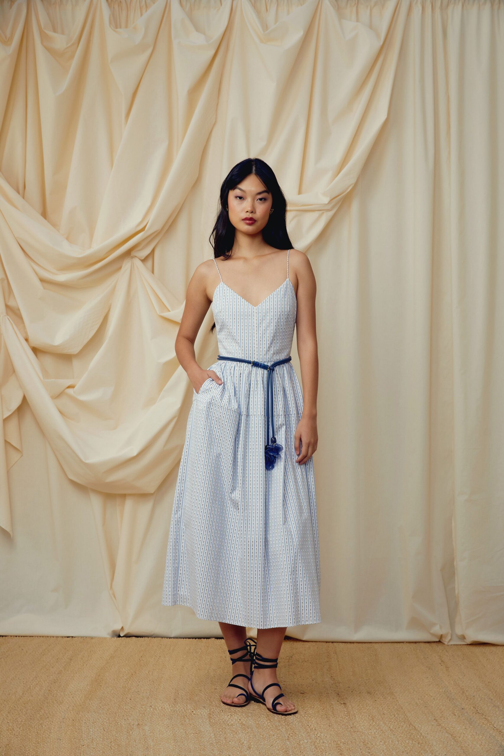 White midi sundress in narrow blue dotted stripes, seamed low on the skirt. Cobalt cord belt knotted loose with a tasseled end at the hip. Flat navy lace-up sandals winding high around the ankle. For walking back from the pensione bakery before the stone streets heat up.