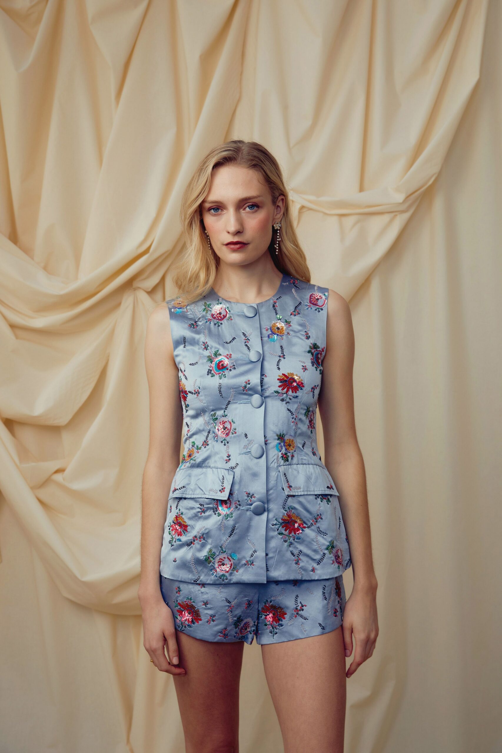 Powder-blue satin vest embroidered with scattered florals and oversized covered buttons. Matching satin shorts, floral embroidery running clean across the side seam. Crystal drop earrings in a narrow chandelier line. Garden wedding brunch while the tablecloths are still being shaken out.