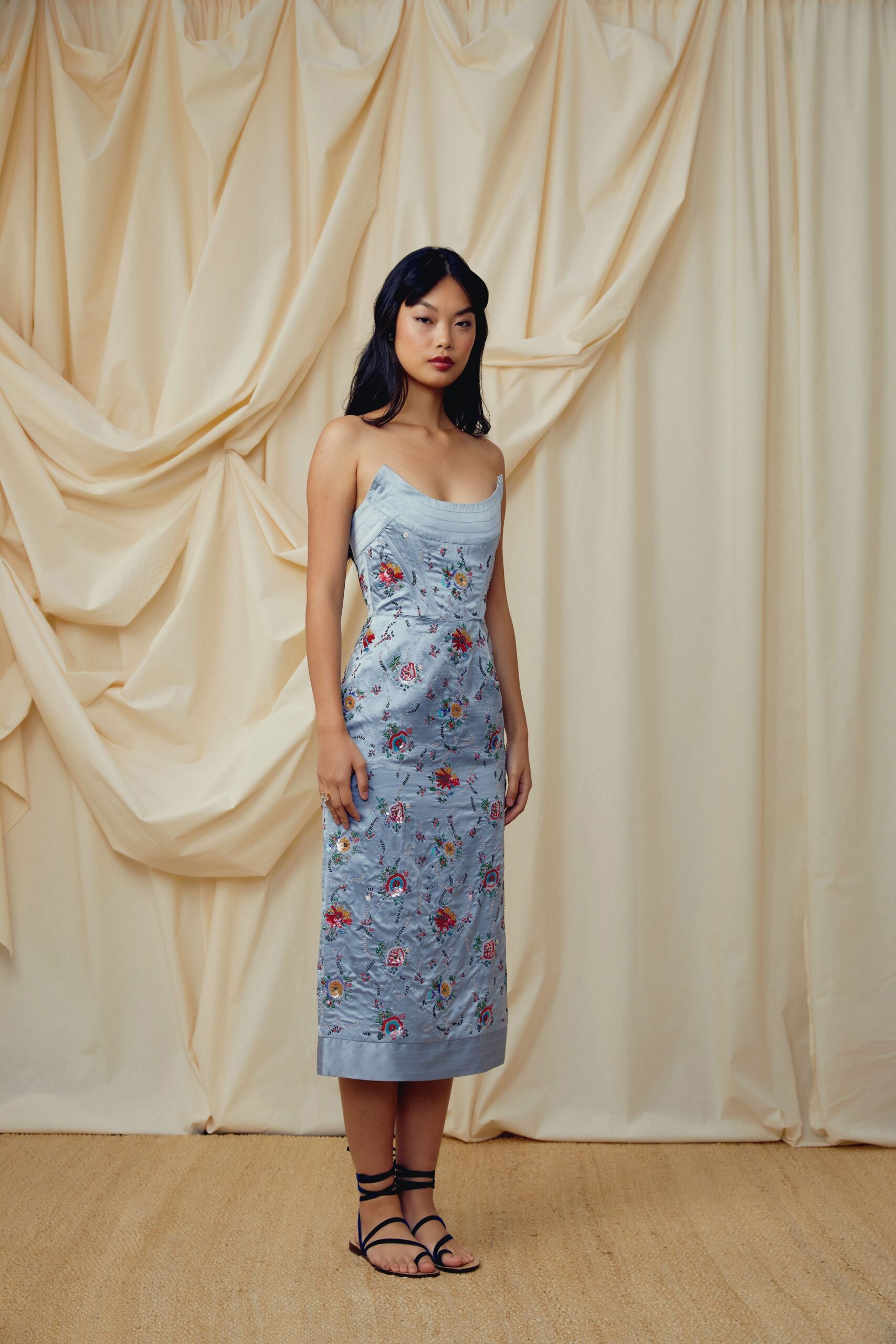 Powder-blue satin column dress with floral embroidery and curved seamed bust. Flat black lace-up sandals, cords winding high at the ankle. Garden wedding spillover, champagne gone warm on the terrace.