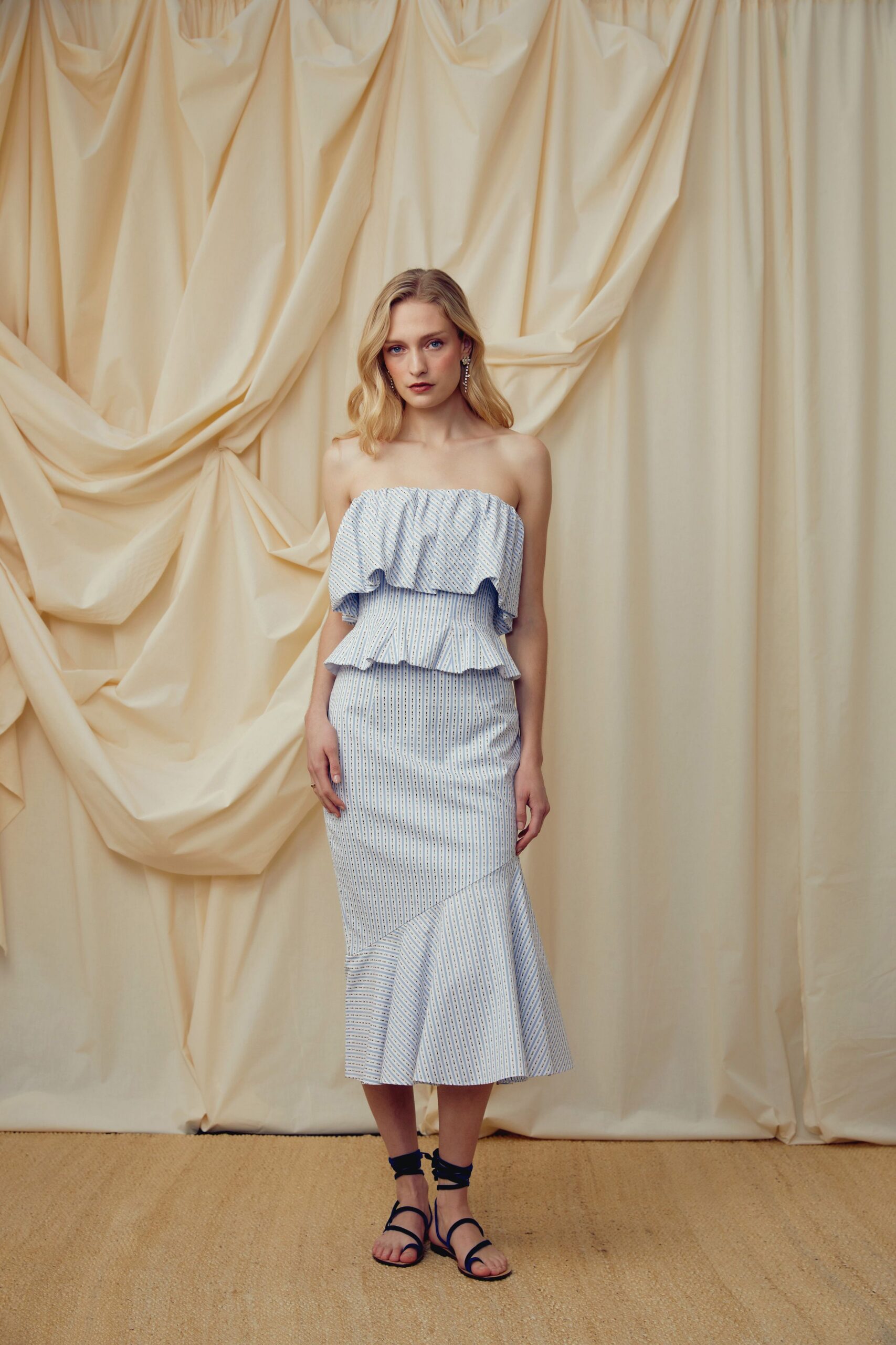 Strapless pale blue pinstripe dress with a ruffled bustier flounce and peplum. Black wraparound sandals, ties crossing the ankle in thin cords. Crystal drop earrings brushing the jaw. Garden wedding, the cake already listing in the heat.