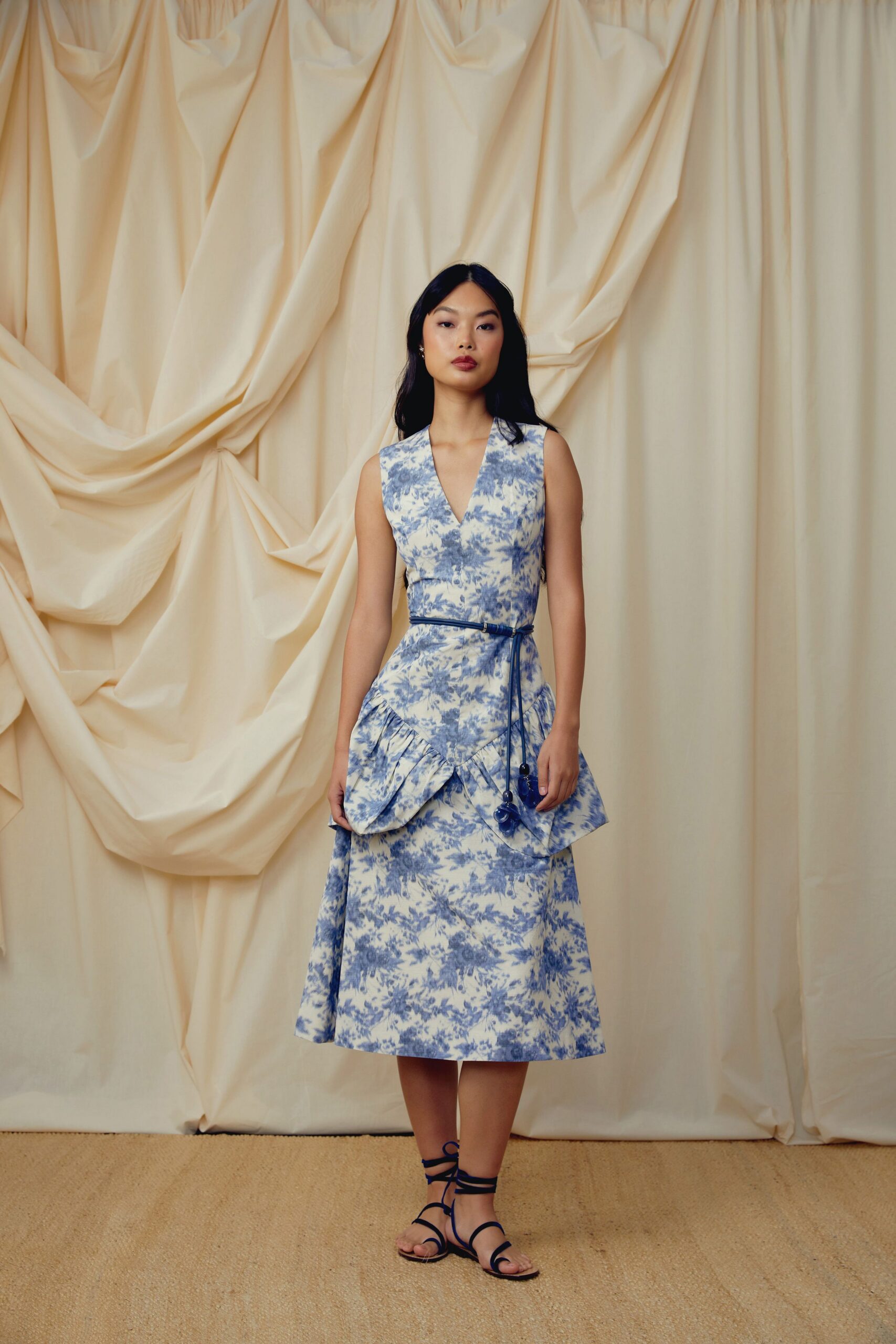 Blue toile sundress with a deep V and petal-shaped peplum flounce. Skinny cobalt cord belt, long tasselled ends weighted with glossy beads. Flat lace-up sandals in inky blue leather. Garden wedding, cake table already listing in the heat.