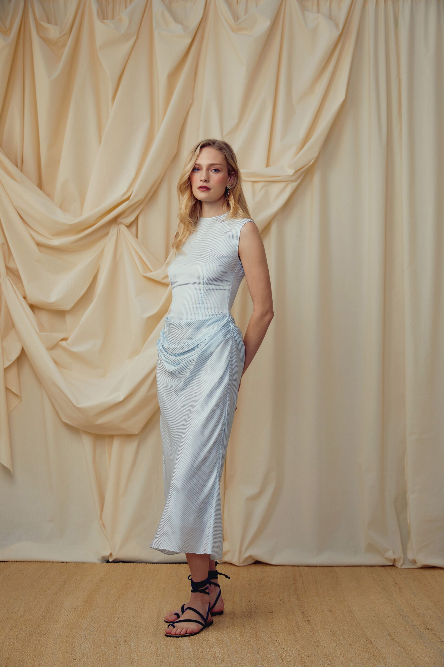 Ice-blue ribbed dress with a bateau neck and gathered hip drape. Black wrap sandals, cords tied high at the ankle. Walking barefoot home from a garden wedding, heels already in your hand.