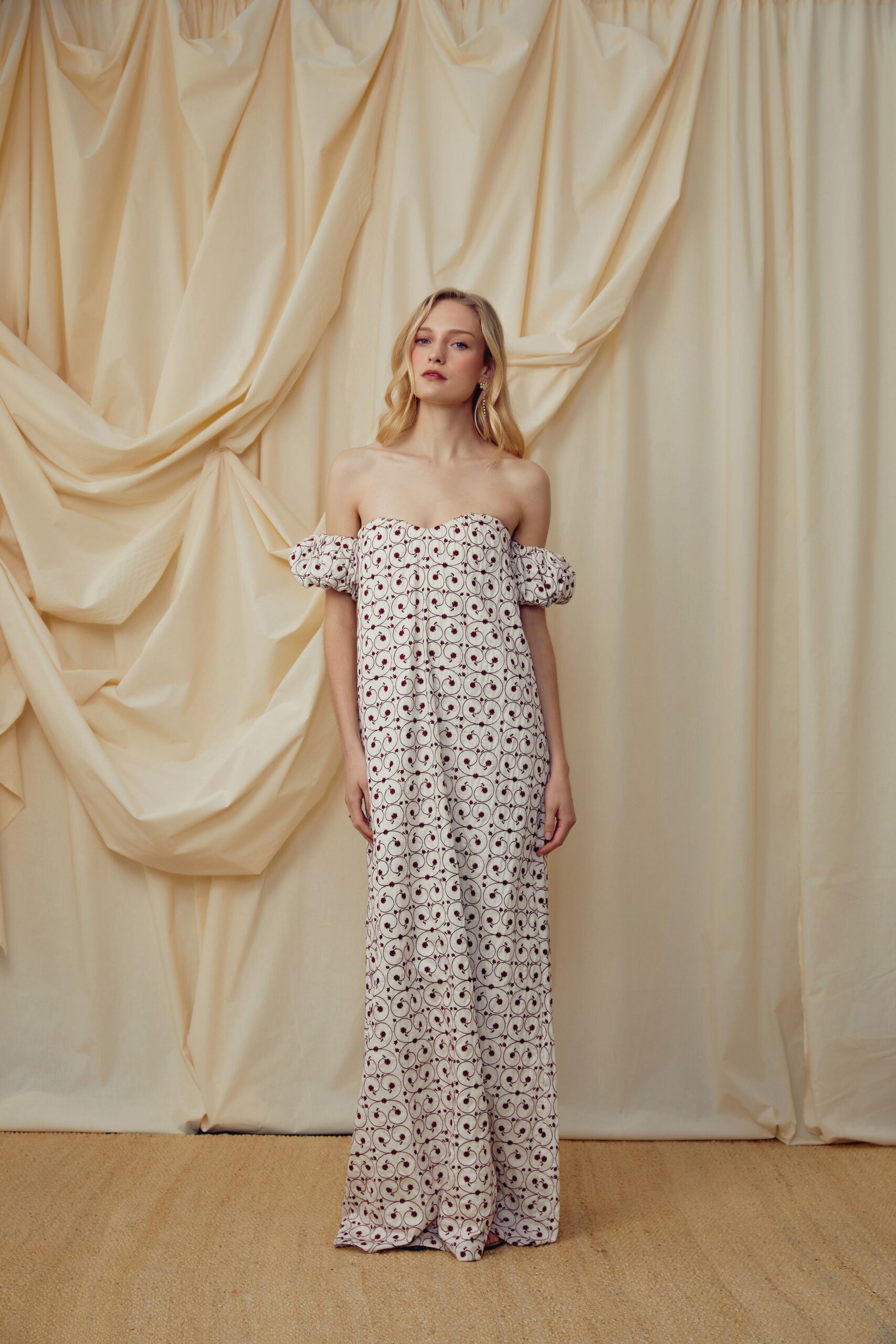 Ivory strapless column gown with puffed off-shoulder sleeves and burgundy vine embroidery. Long crystal drops brushing the jaw. Hotel ballroom before noon, florists still clipping stems into silver bowls.
