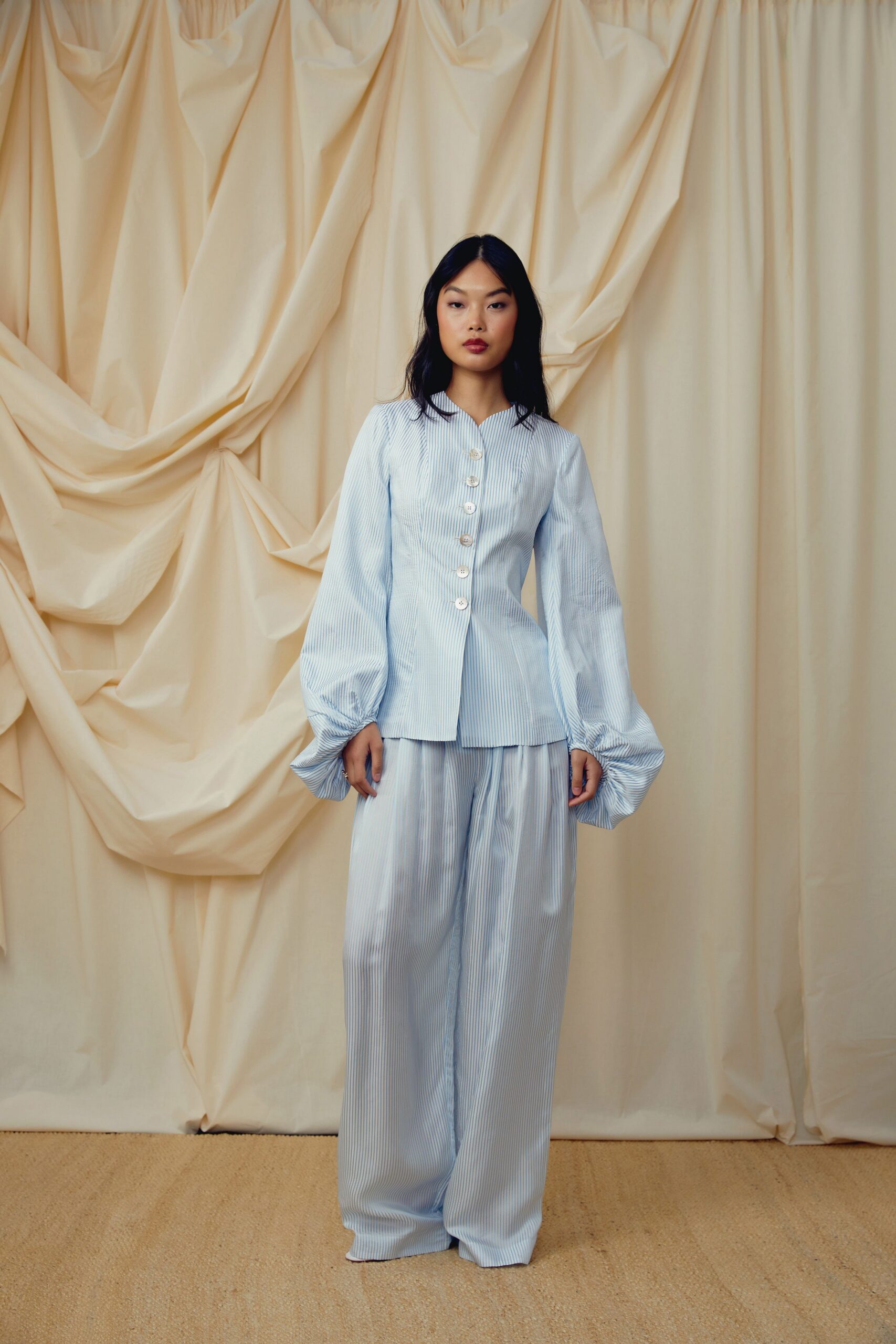 Powder-blue pinstriped blouse, fitted through the waist with gathered balloon cuffs. Matching wide-leg trousers in the same fine stripe, pooling over the feet. Morning light through rented-house curtains, coffee going cold on the sill.