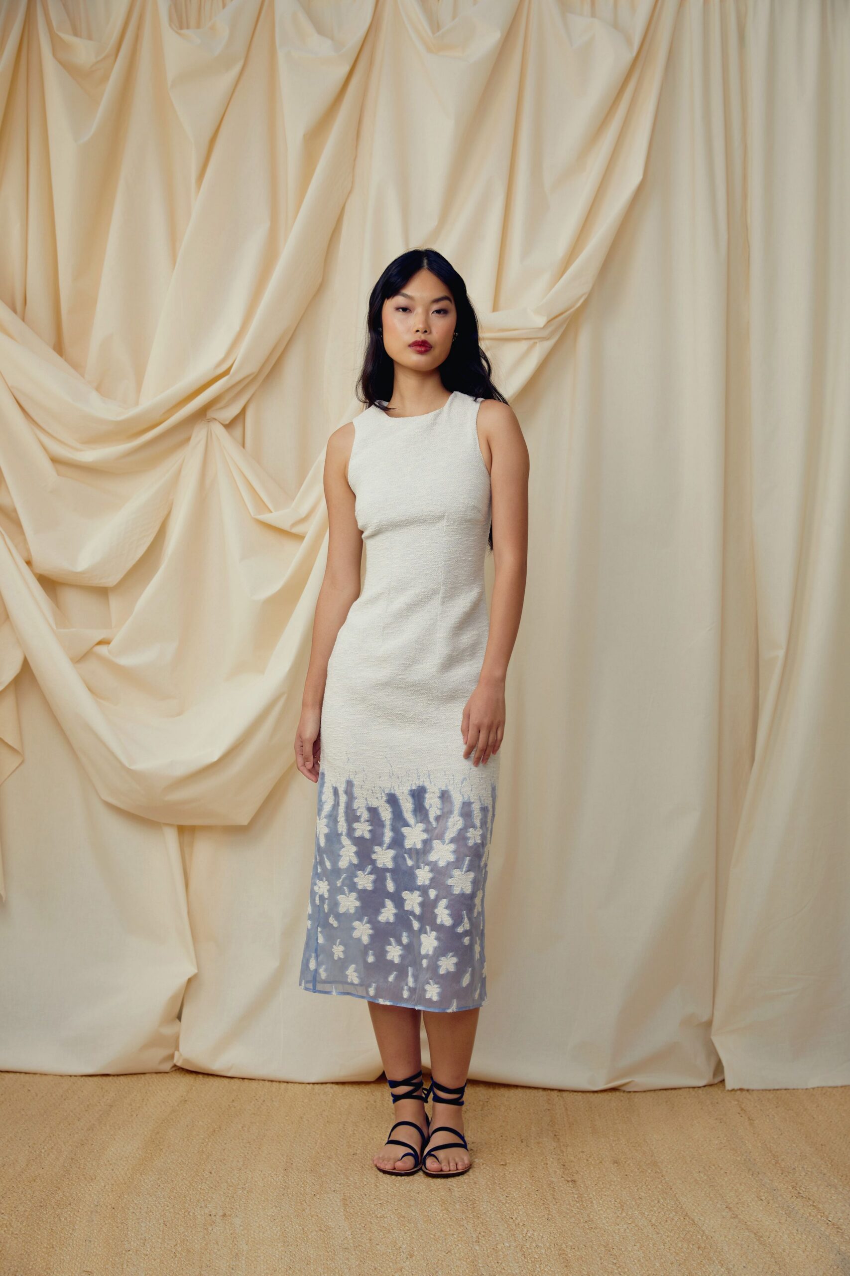Ivory sleeveless column dress in nubby knit, fading into powder-blue floral mesh at the calf. Navy lace-up flat sandals, cords crossing the ankle twice. Garden wedding spillover, standing barefoot-adjacent on the sisal after dusk.