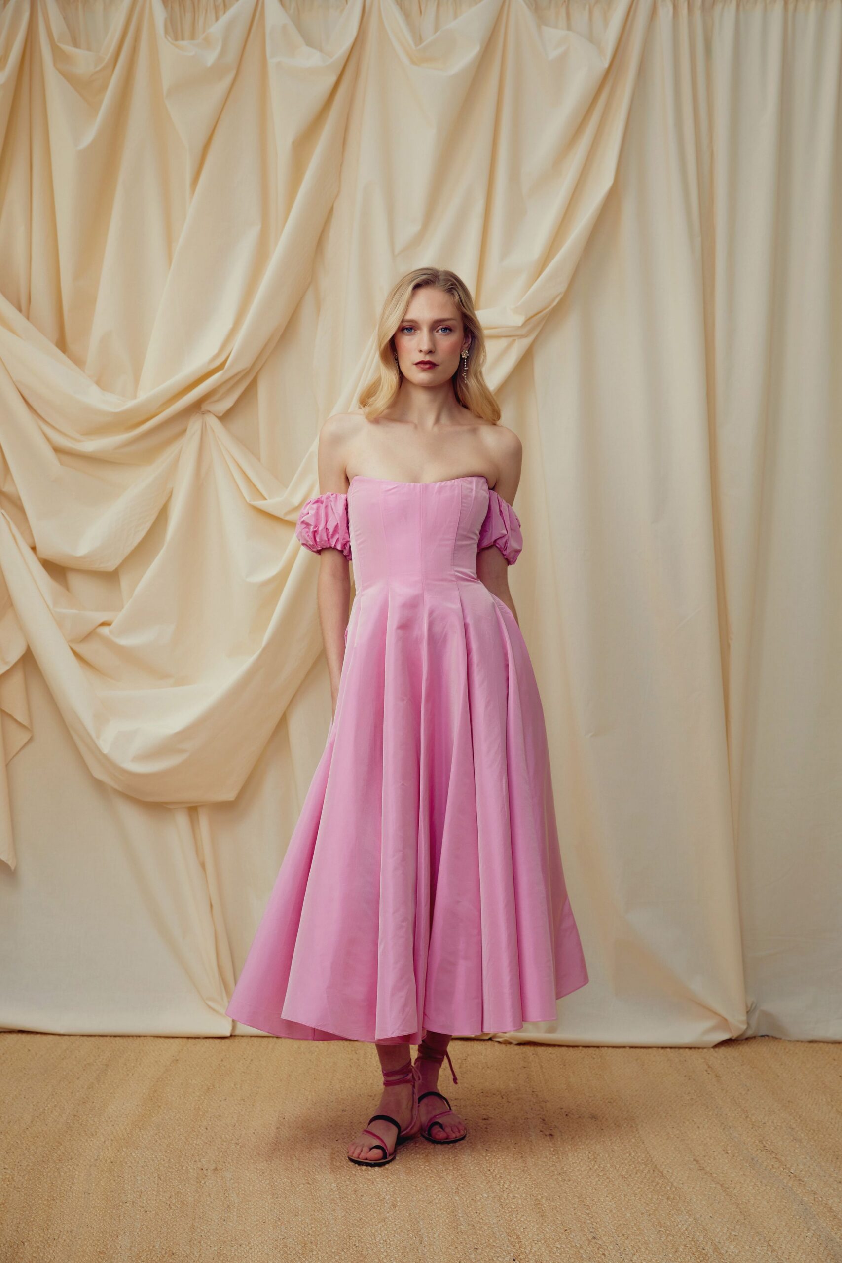Bubblegum pink cotton dress with a boned bodice and dropped puff sleeves. Bare black sandals tied up the ankle with pink cords. Garden wedding, cake table already softening in the heat.