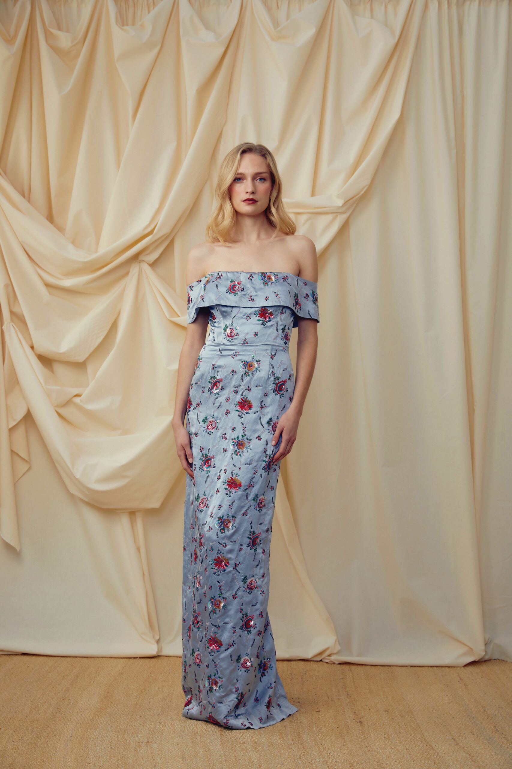 Powder-blue brocade gown with an off-shoulder foldover band and scattered floral embroidery. Garden wedding after the chairs have sunk a little into the lawn.