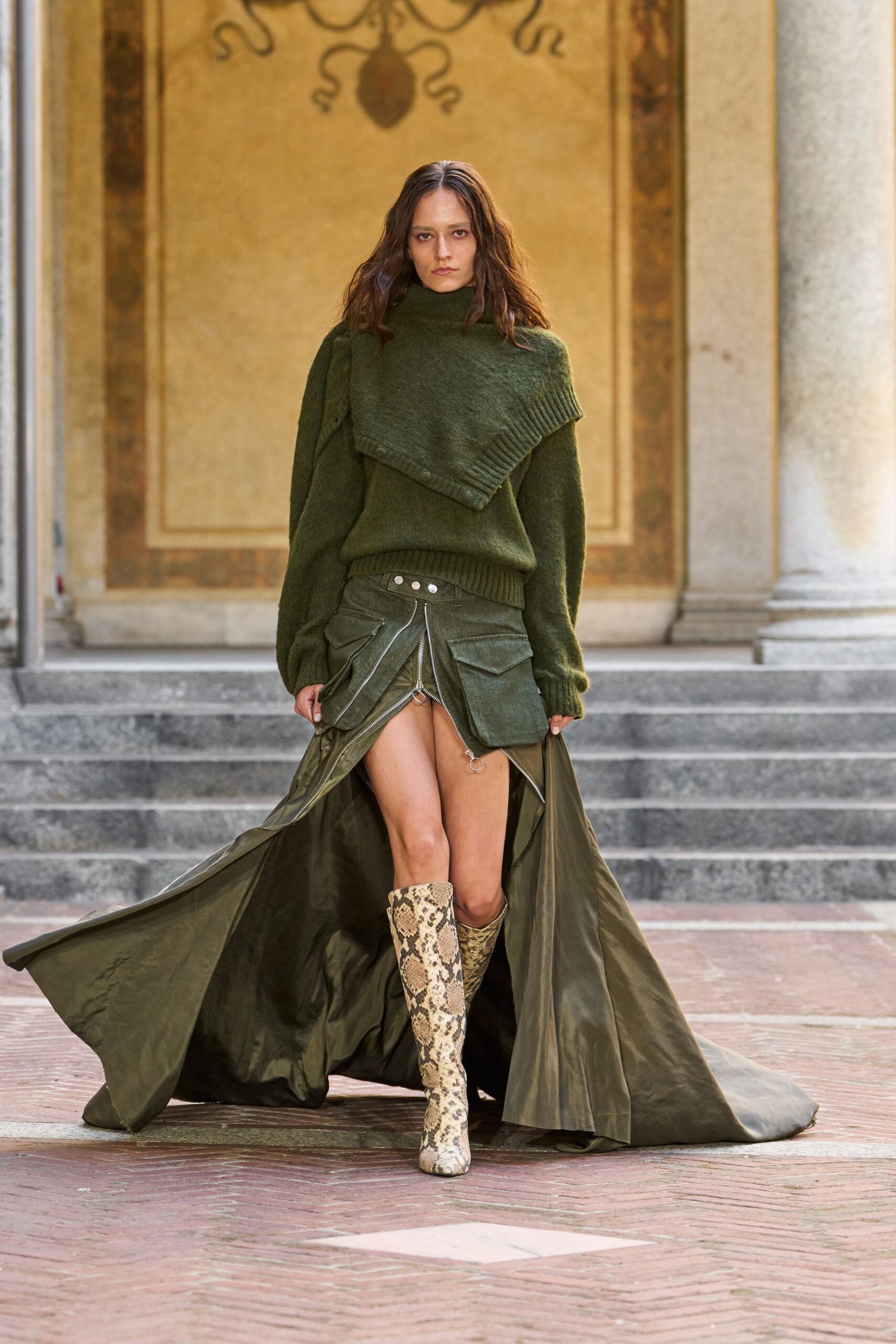 Moss brushed knit sweater with an off-kilter ribbed shoulder cape. Olive utility maxi skirt unzipped to the hip, ring pulls swinging. Snakeskin knee boots in sandy python tones. Crossing a villa courtyard just after rain, train picking up grit.