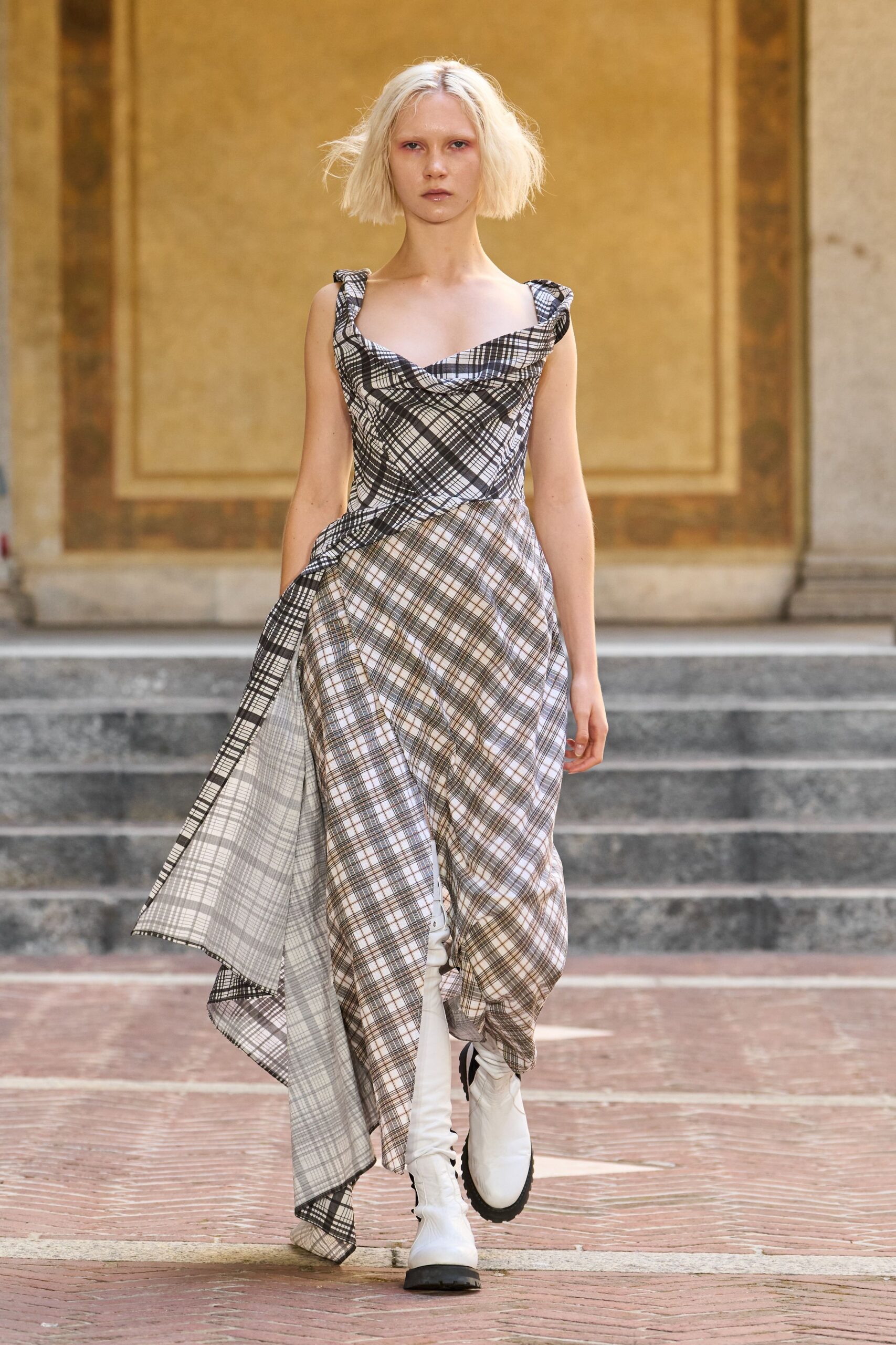 Bias-cut plaid dress with a folded cowl and one long trailing panel. White knee boots with black toe caps and thick lug soles. Crossing a stone courtyard after rain, hem already drinking from the puddles.