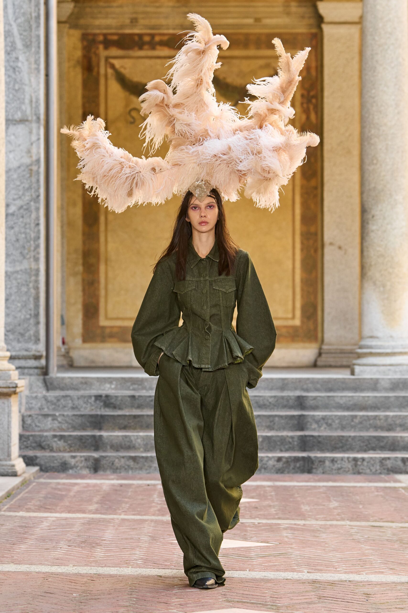 Blush ostrich plumes arcing into antler-like branches above a jeweled brow mount. Moss denim jacket, seamed close through the torso with a fluted peplum. Matching trousers cut very full, breaking into heavy folds over the shoes. Black square-toe shoes. Crossing a frescoed courtyard as the wedding guests finally drift outside.