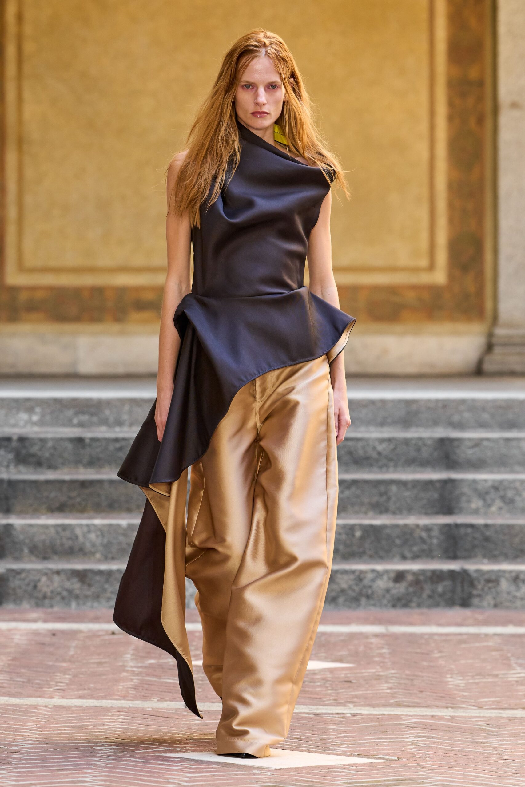 Ink satin top cut on the bias, neck collapsing into a cowl. Asymmetric flounce jutting from the hip, pale reverse flashing underneath. Champagne satin trousers with a soft break and creased, pouching front. Crossing a frescoed courtyard just before the museum doors open.