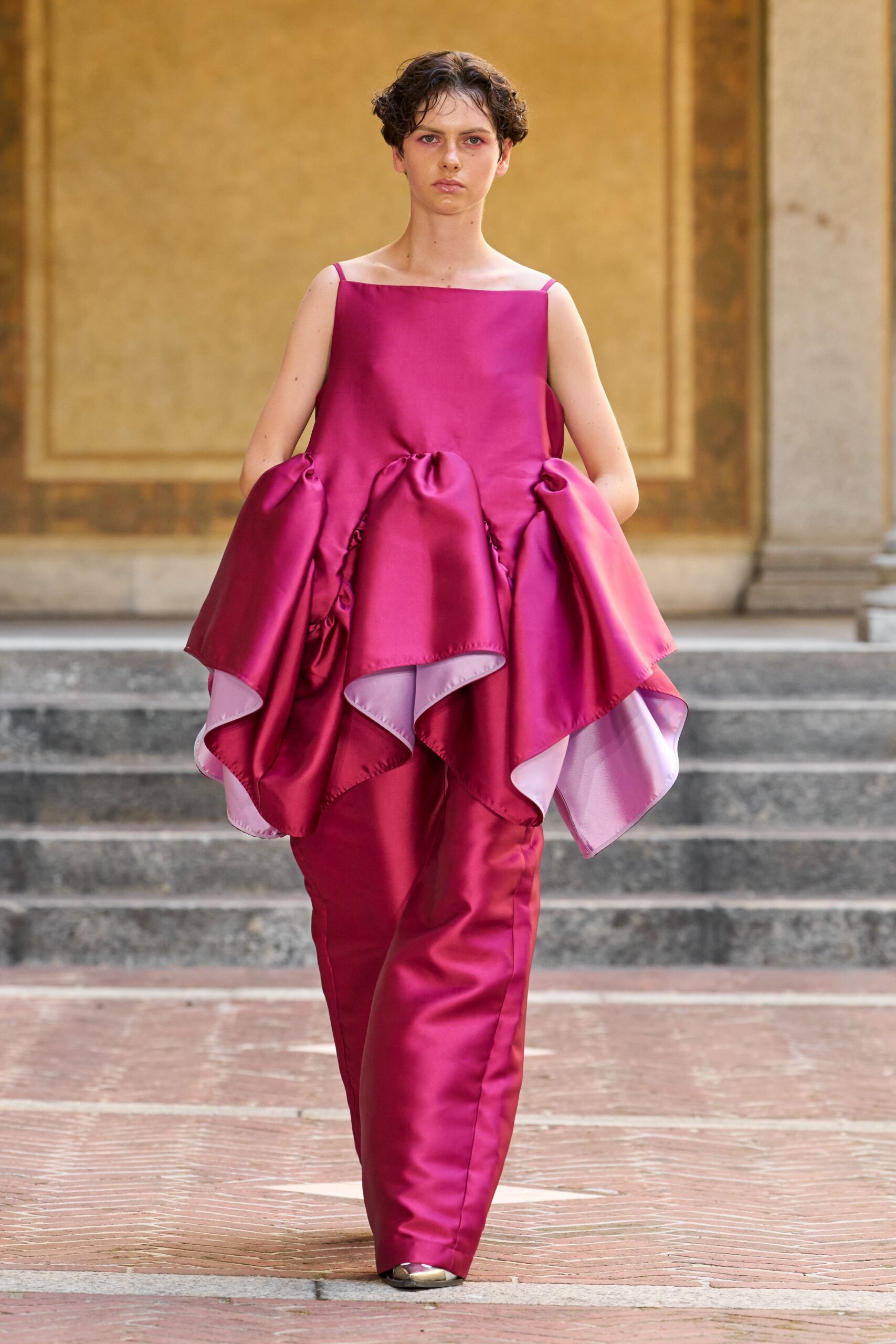 Fuchsia duchesse satin shell, square neckline on thin straps. Ballooned overskirt folded into deep scallops, pale pink underside flashing. Matching satin trousers falling straight with a soft break at the shoe. Metallic almond-toe pumps. Crossing a frescoed courtyard after the wedding crowd has gone inside.
