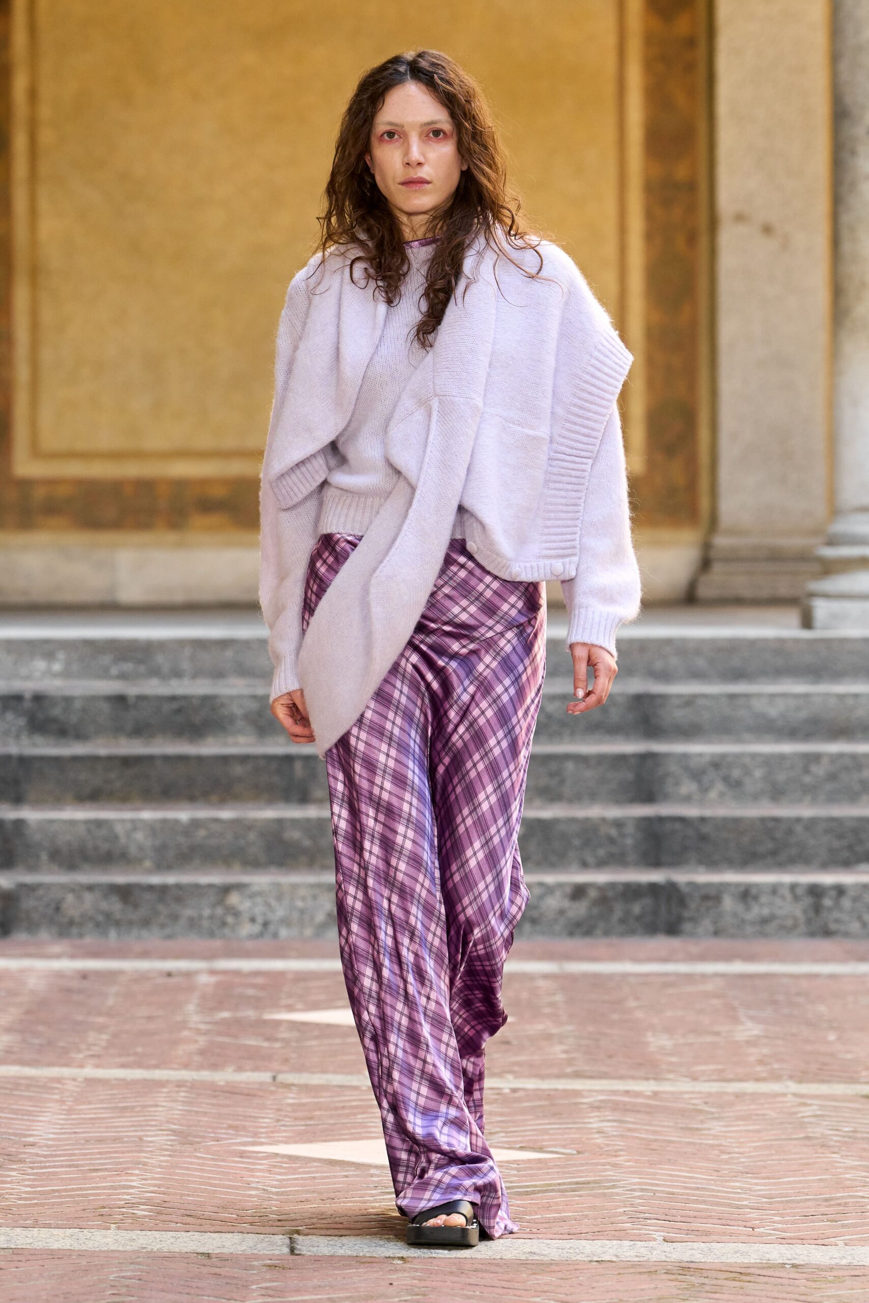 Pale lilac fuzzy knit sweater with a thick ribbed hem. Matching brushed knit scarf draped broad across the shoulders. Bias-cut satin trousers in mauve plaid, pooling over the sandal. Black platform slide sandals. Crossing a museum courtyard before the stone has given up the day's heat.
