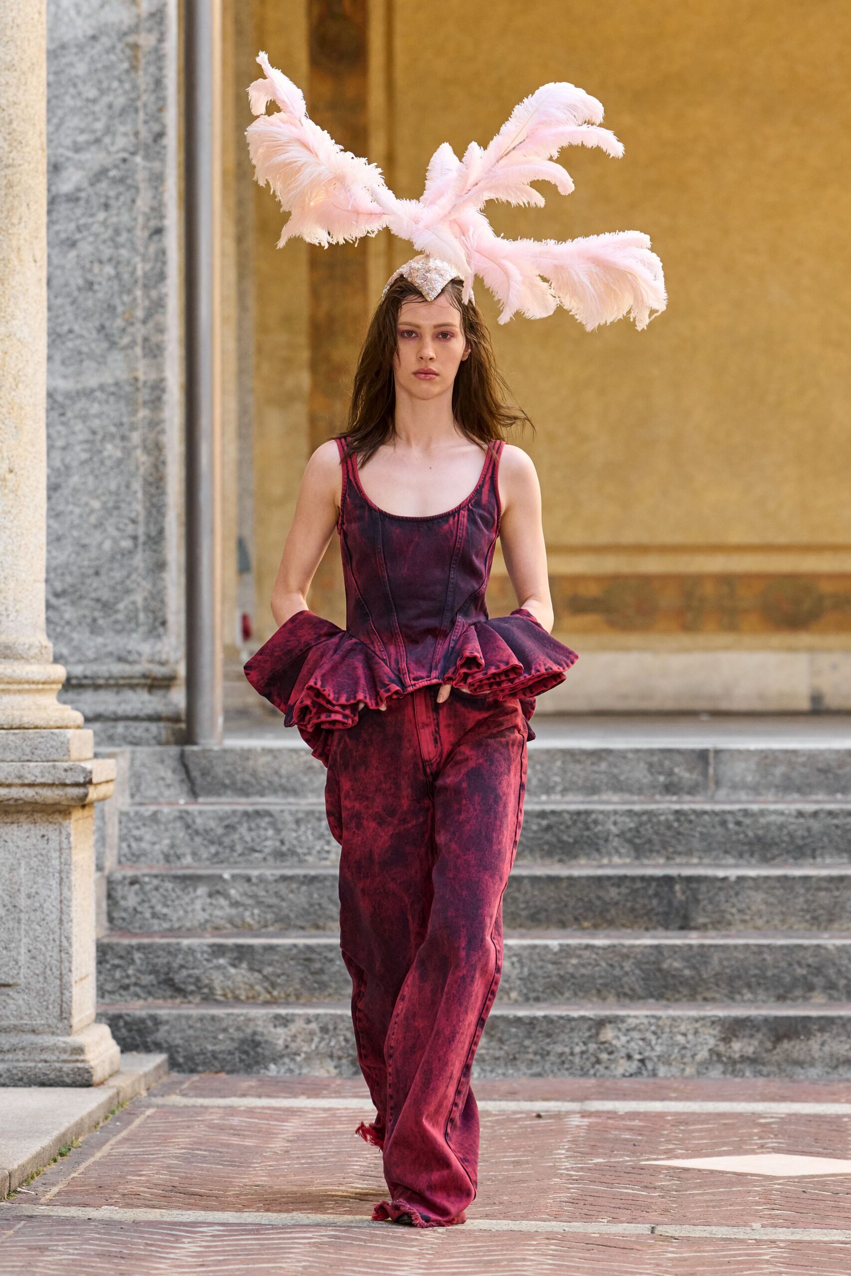 Pale pink ostrich plumes bursting from a silver skullcap. Acid-washed claret corset top, peplum flouncing out in deep ruffles. Matching low-slung jeans, hems dragging into frayed tassels. Crossing a sunstruck cloister courtyard after the costume ball ran late.