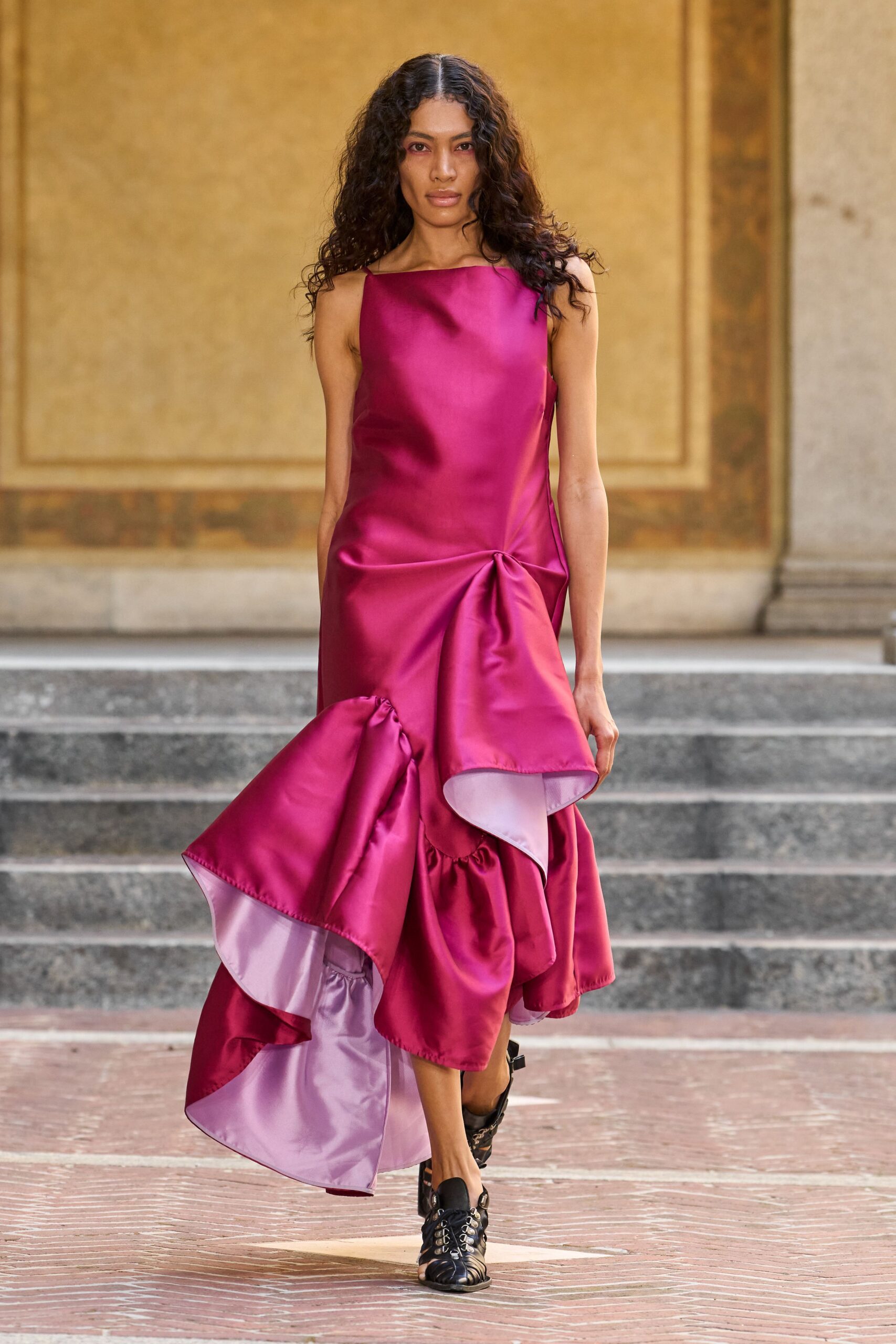 Fuchsia duchesse satin column dress, hip pinched into a spill of ruffled panels. Black cutout lace-up flats with silver eyelets. Crossing a sunstruck courtyard after the wedding guests have drifted inside.