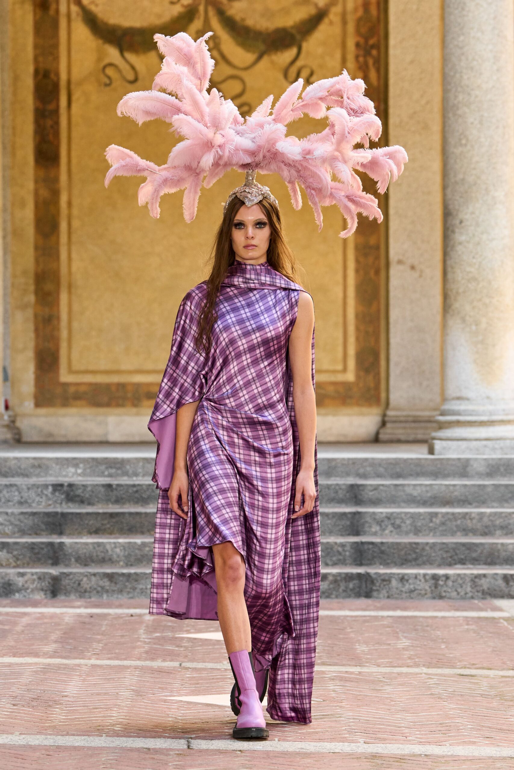 Pale pink ostrich plumes flaring from a crystal-capped headband. Asymmetric mauve plaid dress in liquid satin, scarf neck and cascading panel. Lilac ankle boots with thick black lug soles. Crossing a villa courtyard after midnight, still wearing the party centerpiece.