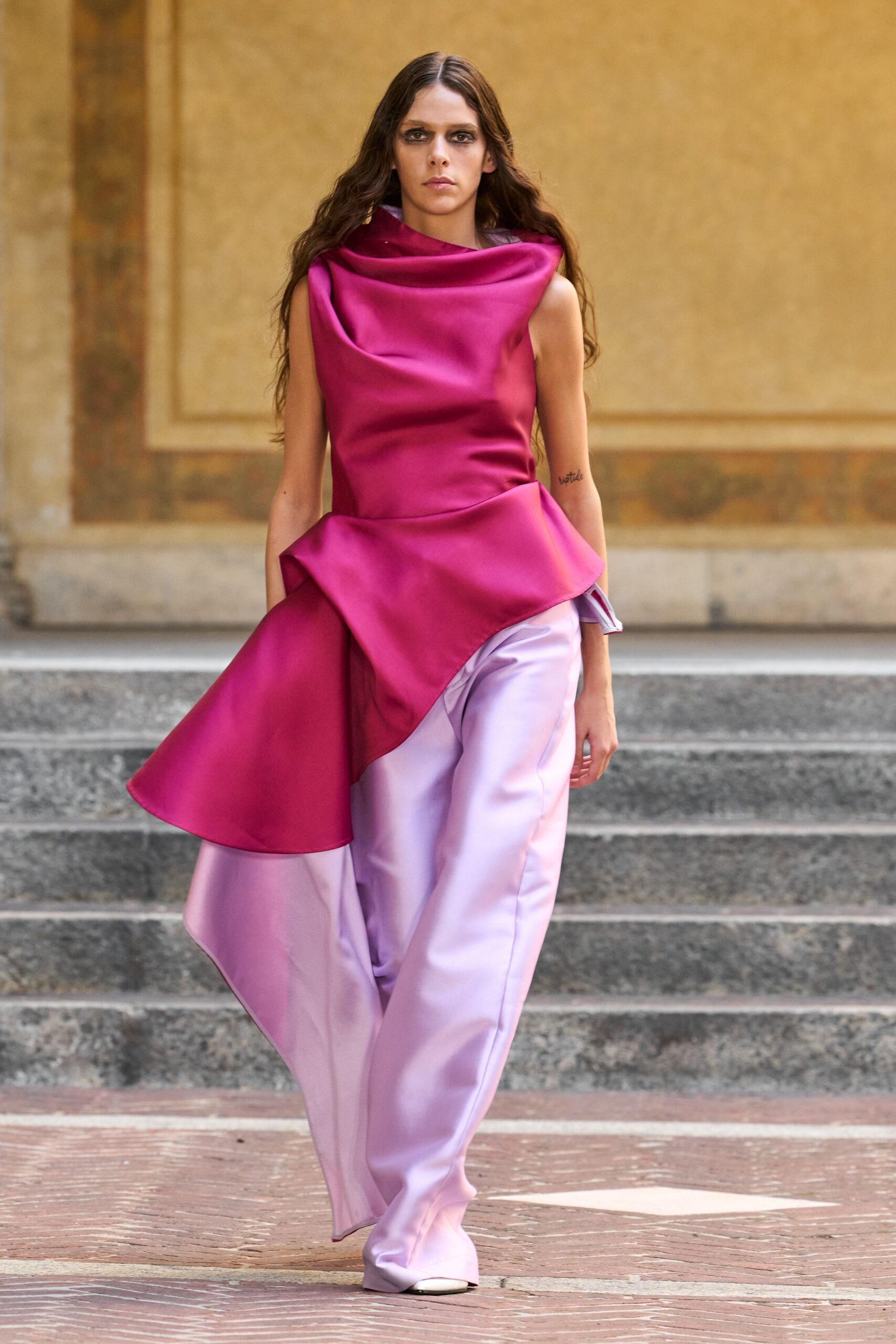 Fuchsia duchess satin top with a deep draped collar and sculpted peplum flare. Pale lilac satin wide-legs, pooling over the shoe with a sharp front crease. White pointed toes just breaking the hem. Crossing a palace courtyard after rain, carrying the speech in your head.