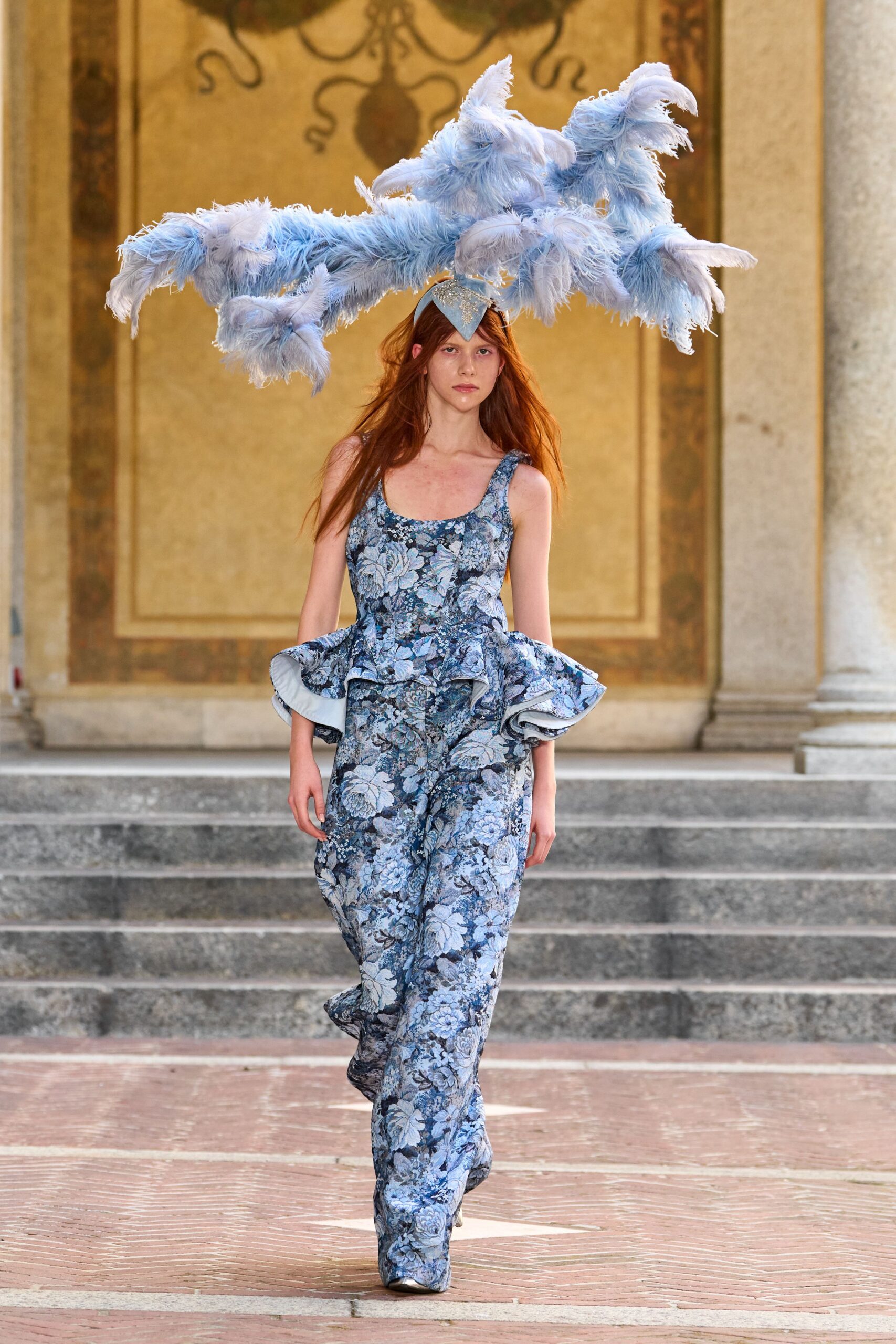 Powder-blue plumes branching wide from a glittered cap. Blue floral brocade jumpsuit with a low scoop neck. Stiff hip flounces, pale lining flashing under each curl. Silver pointed toes just breaking the trouser hem. Crossing a palace loggia while the string quartet is still tuning.