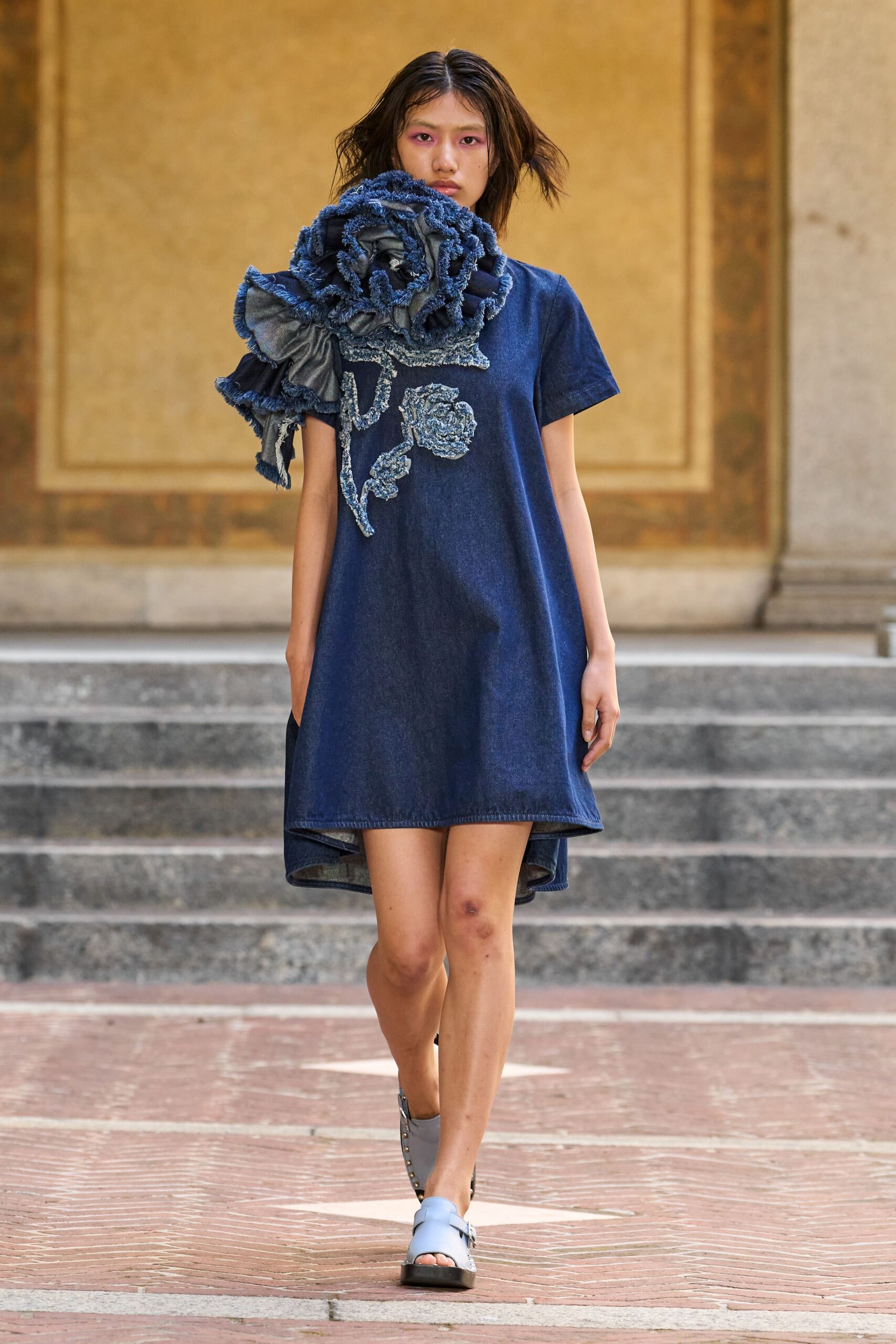 Oversized indigo denim rose scarf, frayed petals swallowing one shoulder. Indigo chambray T-shirt dress with a boucle rose trailing down the chest. Pale blue leather slides studded along the black sole. Crossing a sunstruck cloister courtyard with the bouquet still pinned to you.