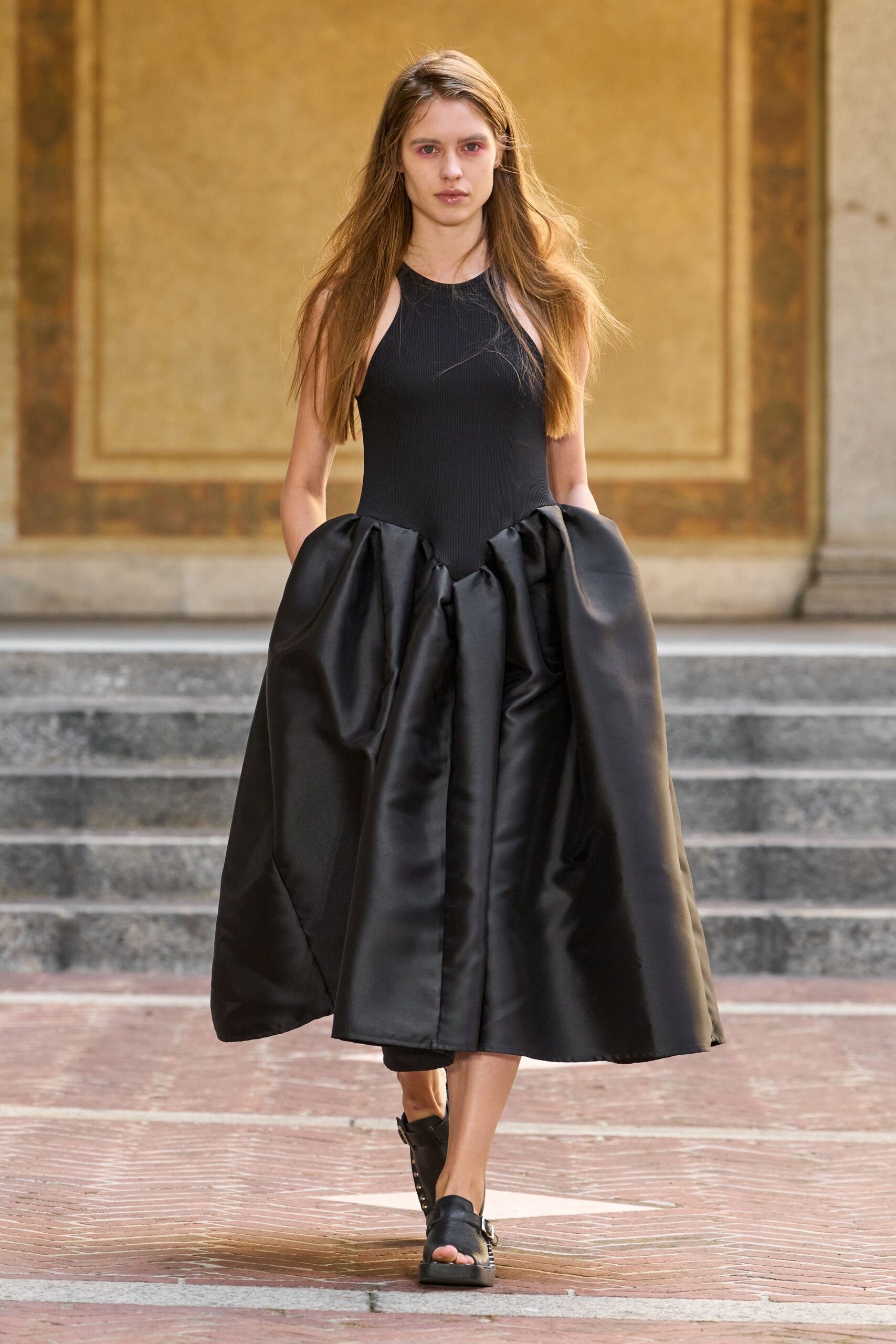 Black ribbed jersey bodice cut to a halter line. Voluminous black satin skirt, deep pleats bursting from a dropped waist. Black leather sandals with a side buckle and split toe opening. Crossing a sunstruck cloister courtyard after the bells have finished.