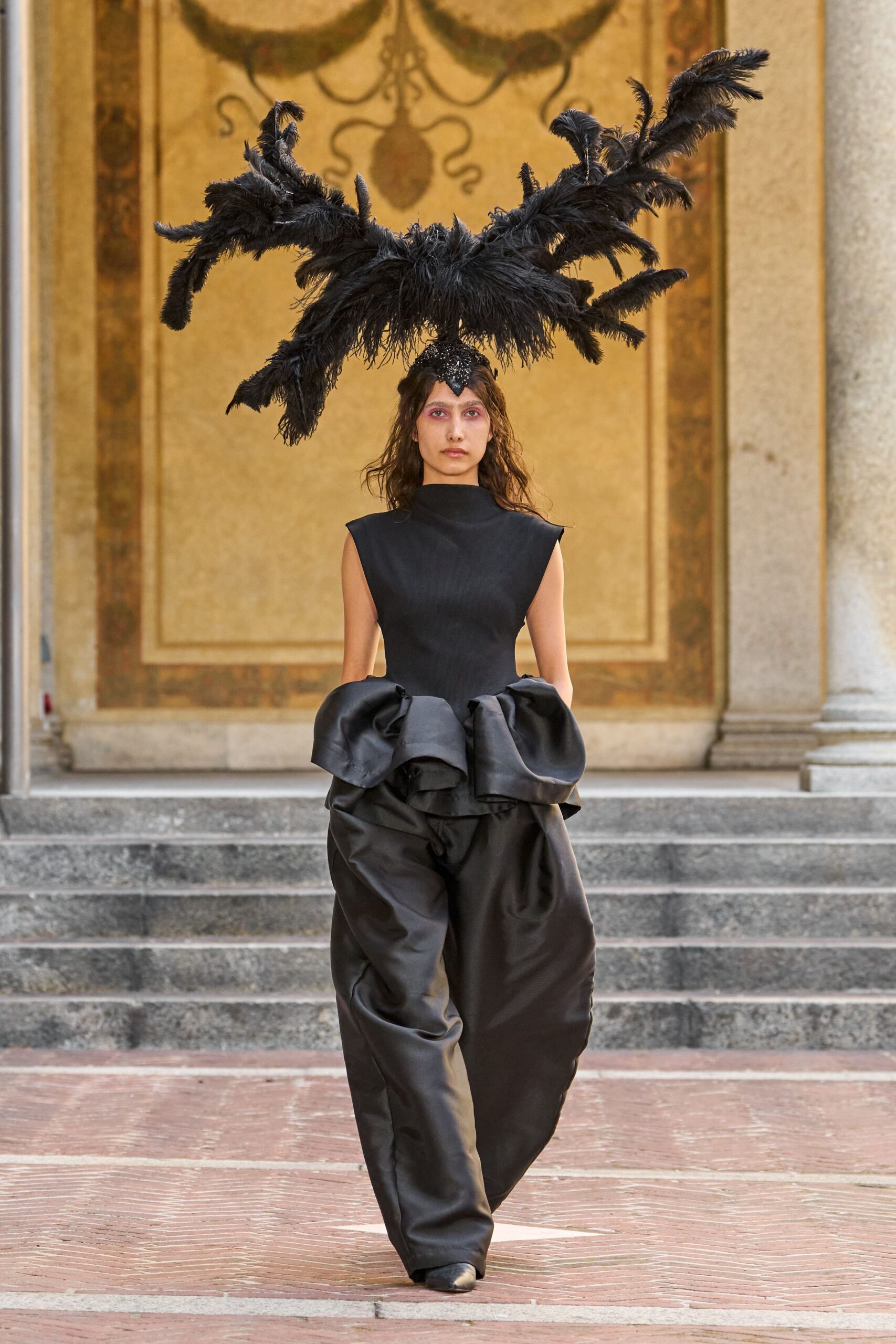 Black ostrich plumes arcing wide from a beaded skullcap. Black sleeveless top with a high funnel neck and sharp shoulders. Black satin balloon trousers, caught by oversized bows at the hips. Pointed black leather boots peeking from the pooled hem. Crossing a frescoed courtyard just before the wedding guests arrive.