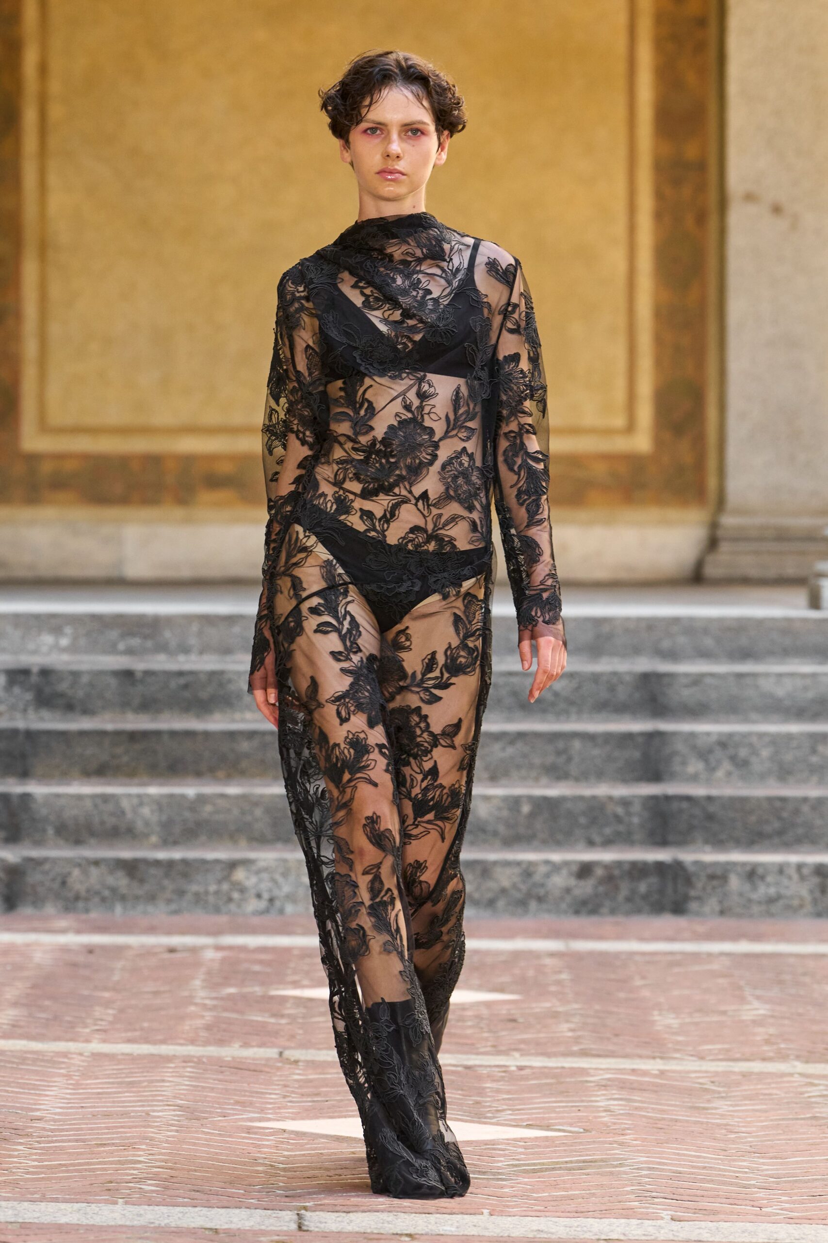 Black floral corded lace over nude tulle, one strap slipping off the shoulder. Black triangle bralette visible beneath the lace. High-cut black briefs under the transparent skirt. Pointed boots swallowed by the trailing lace hem. Crossing a frescoed courtyard before the rain reaches the stone steps.